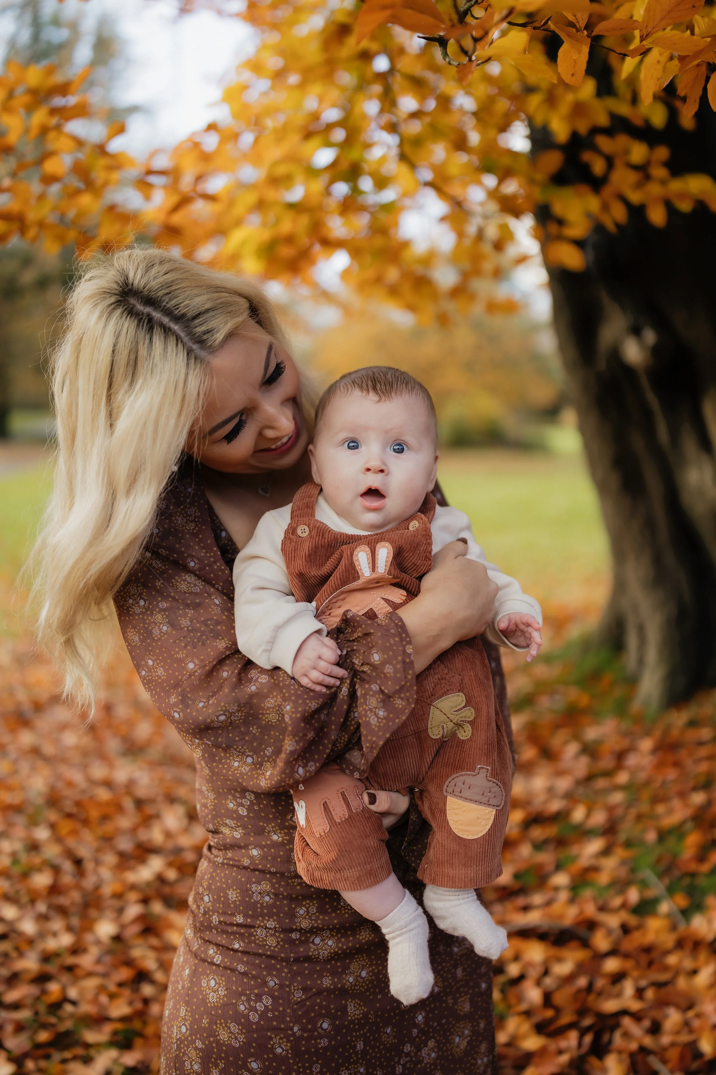 A woman with long blonde hair holds a surprised baby with blue eyes outdoors during autumn, surrounded by orange and yellow leaves on trees.
