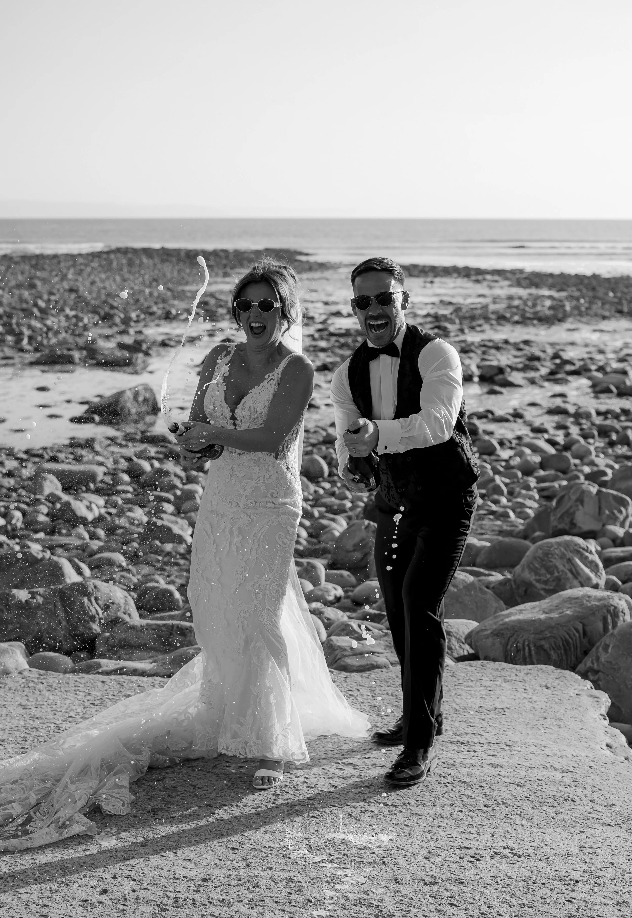 Black and white photo of a bride and groom celebrating on a rocky beach, wearing sunglasses, with the bride in a lace wedding dress and the groom in a tuxedo vest. They are spraying champagne and appear joyful.