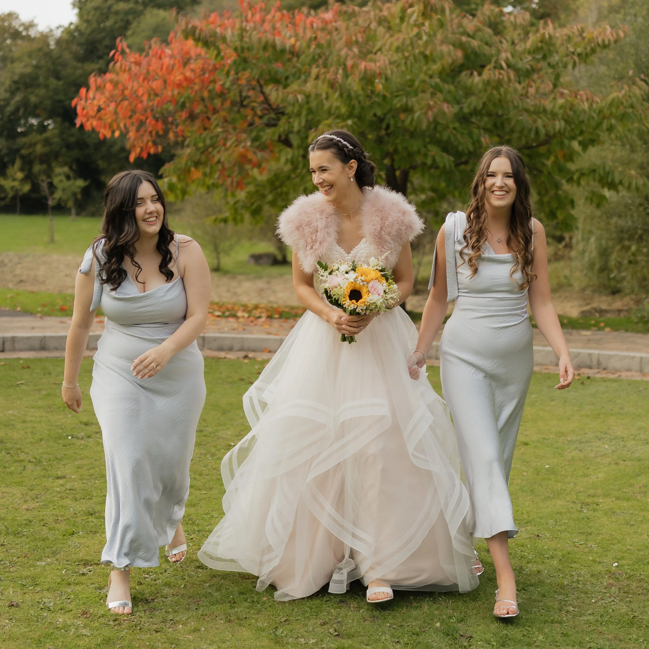 A bride in a white wedding dress with a pink feathered shawl holding a bouquet of sunflowers and roses, walking with two bridesmaids in light gray dresses, on a grassy area with autumn trees in the background.