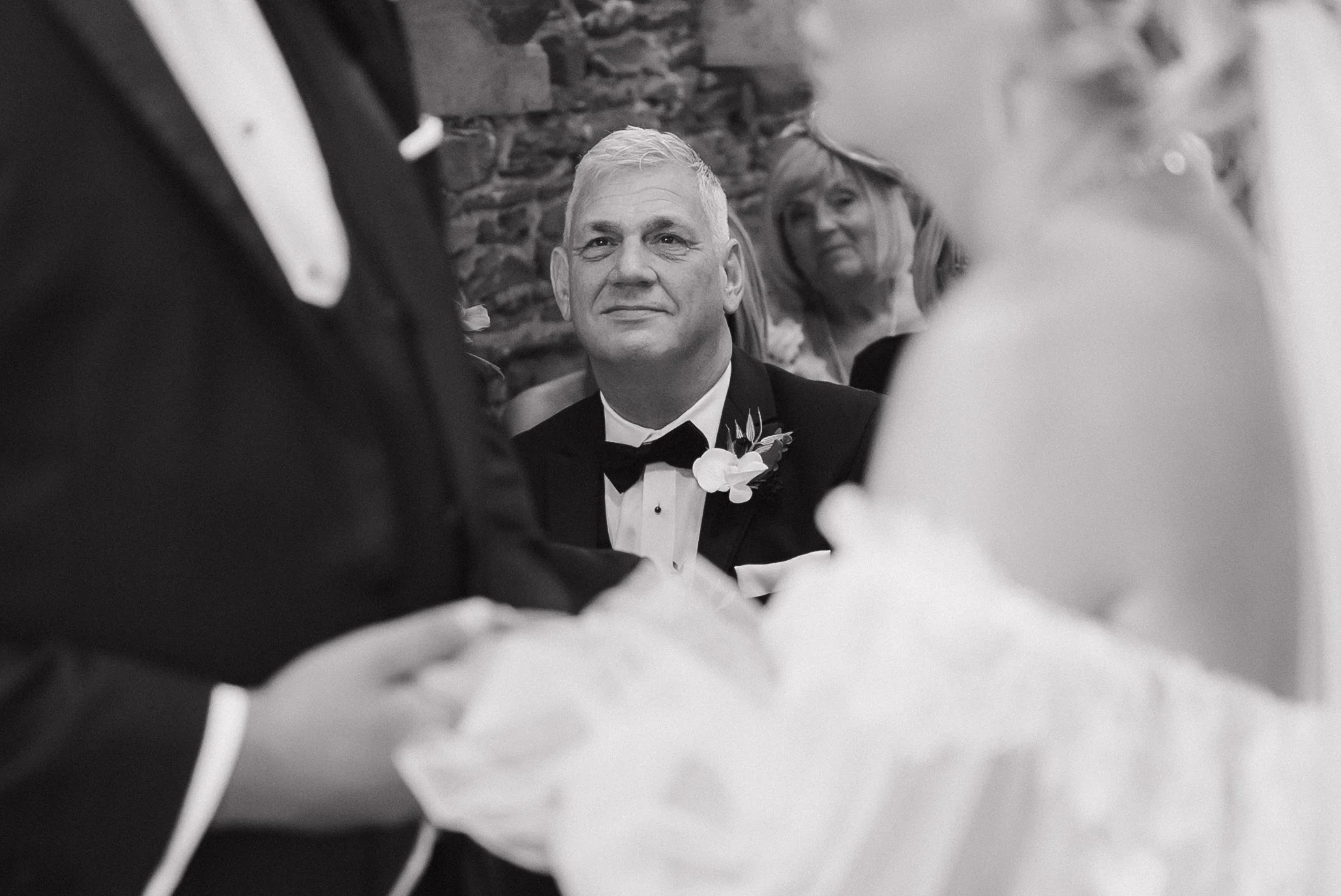 A black-and-white photo of a man in a tuxedo with a bow tie, sitting and looking attentively during a wedding ceremony. A blurred bride and groom hold hands in the foreground, and a woman with glasses and light-colored hair looks on from the backgrou