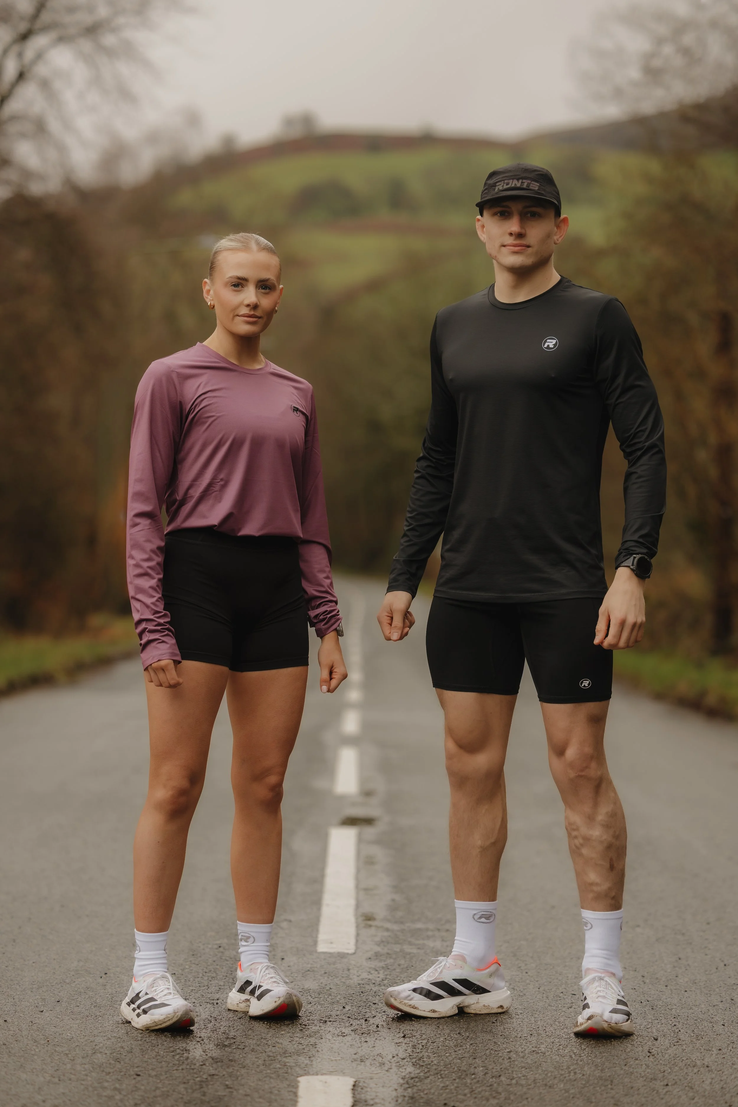 Two runners, a woman and a man, standing on a deserted country road surrounded by trees with fall foliage, dressed in athletic gear ready for running.