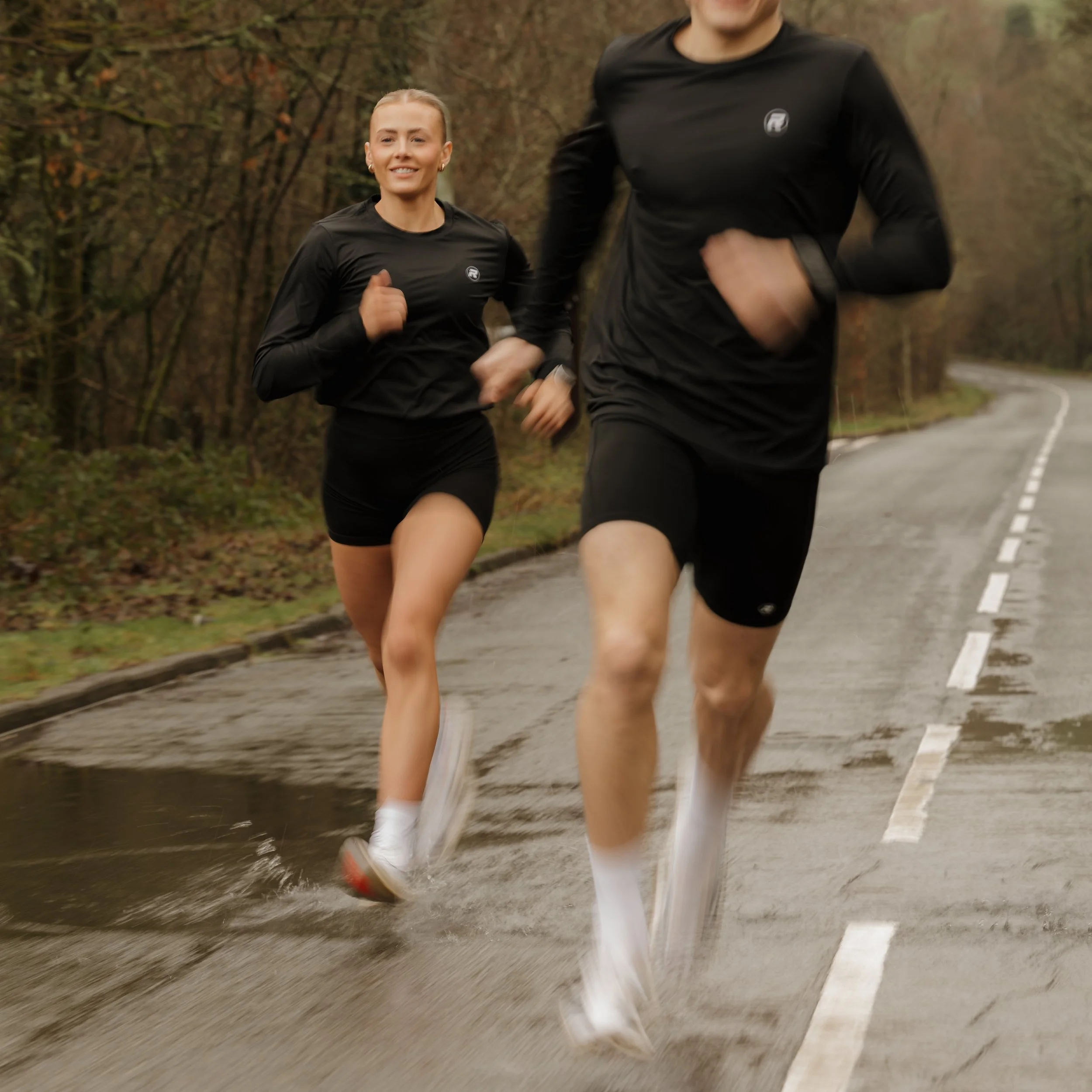 Two people running outdoors on a paved road surrounded by trees. They are wearing black athletic clothing and white running shoes.