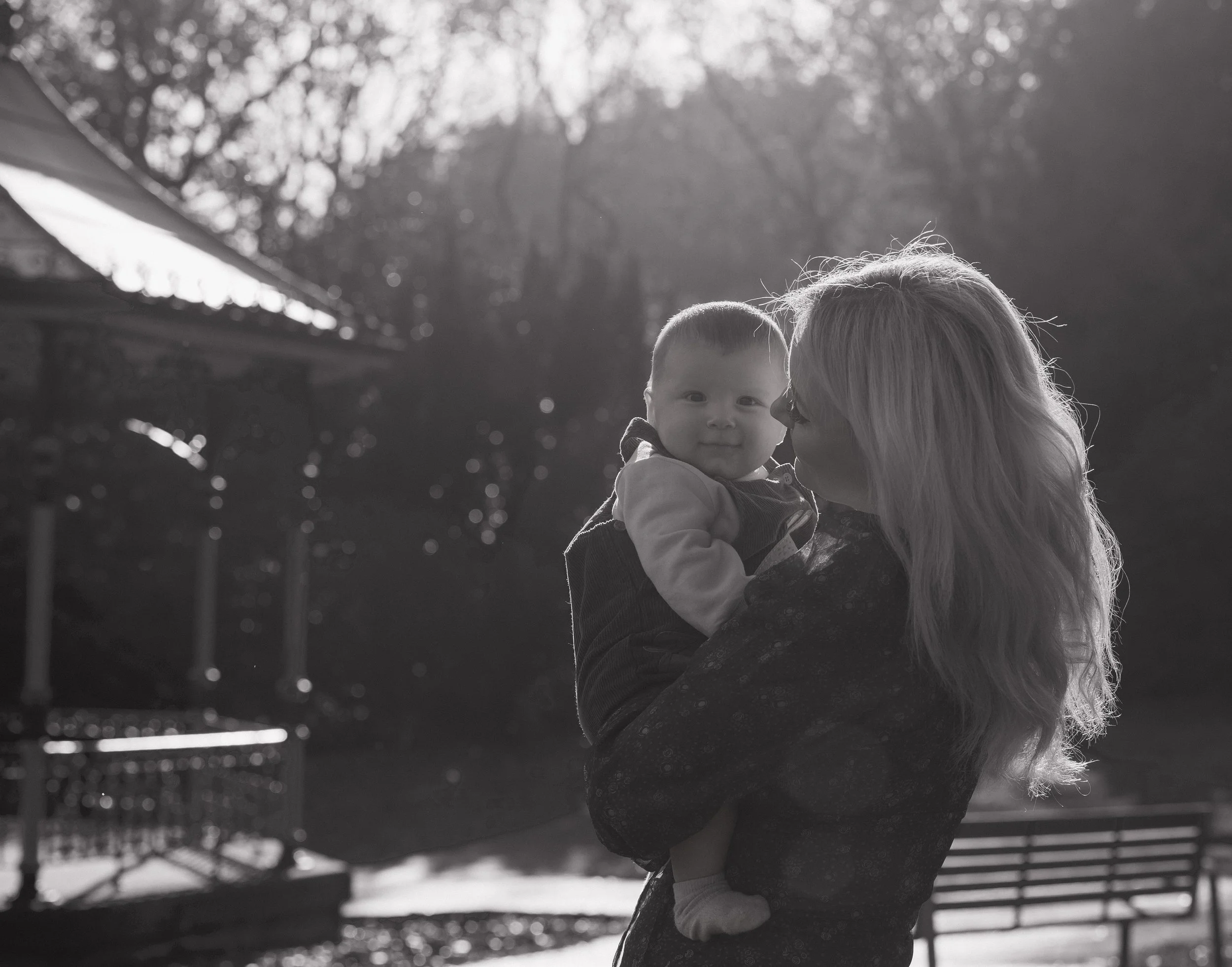 A woman holding a young child outdoors near a park with trees and a pavilion visible in the background, backlit by sunlight.