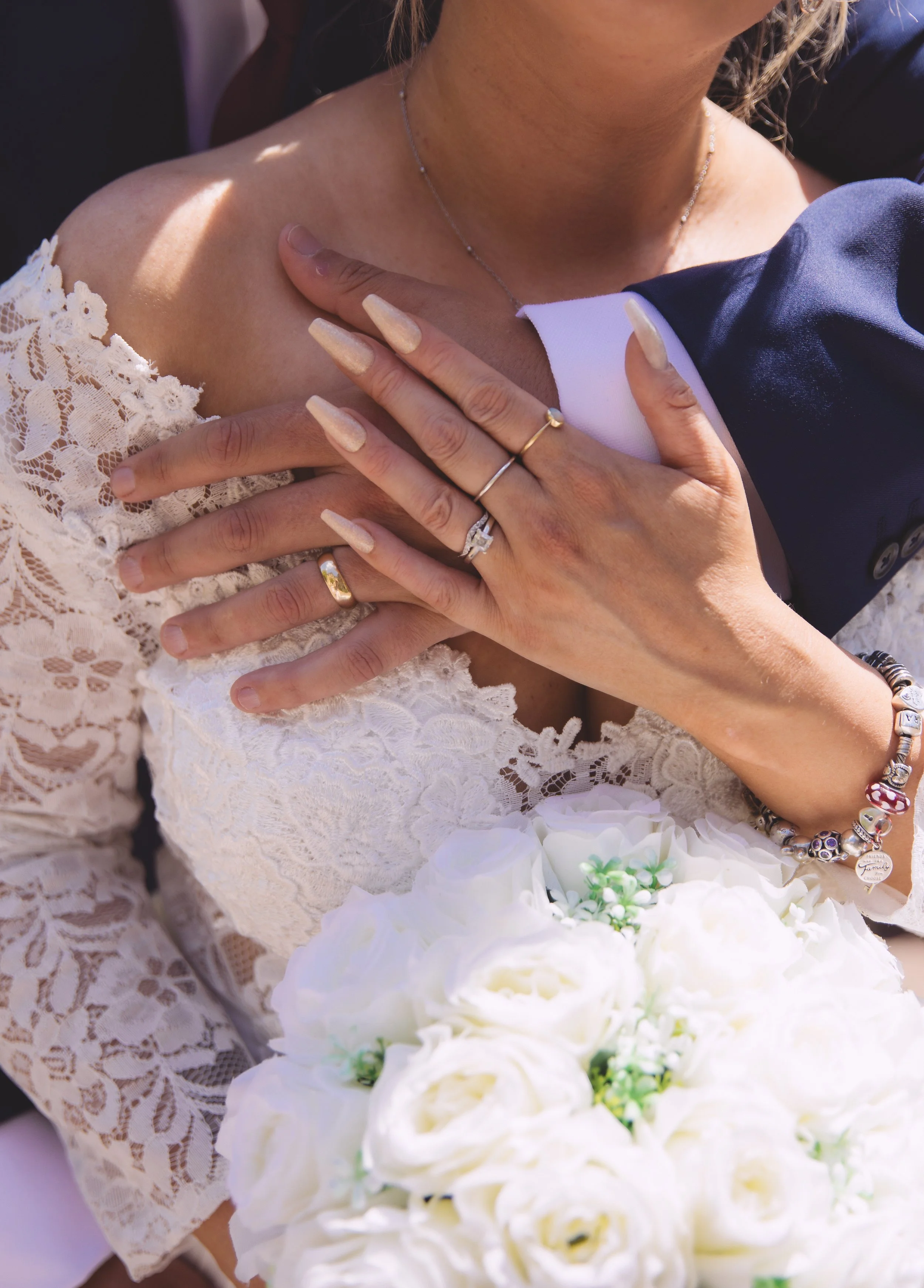 Close-up of a bride in a lace wedding dress holding a bouquet of white roses. Hands with wedding rings are placed on her shoulder, revealing a bracelet and elegant fingernails.