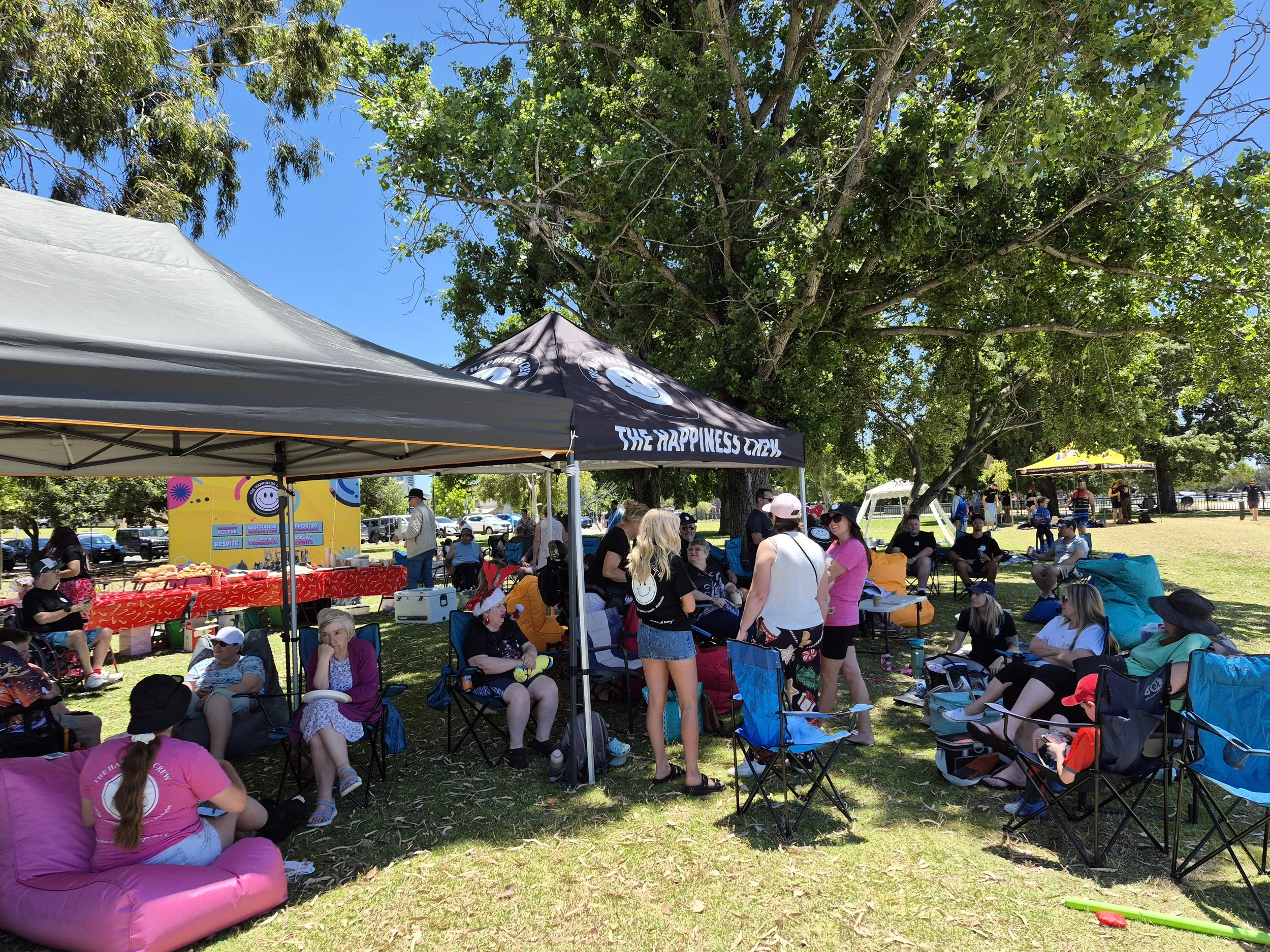 People gathered outdoors under tents and trees at a daytime event, sitting in chairs and on cushions, with tables and colorful decorations, on a sunny day.