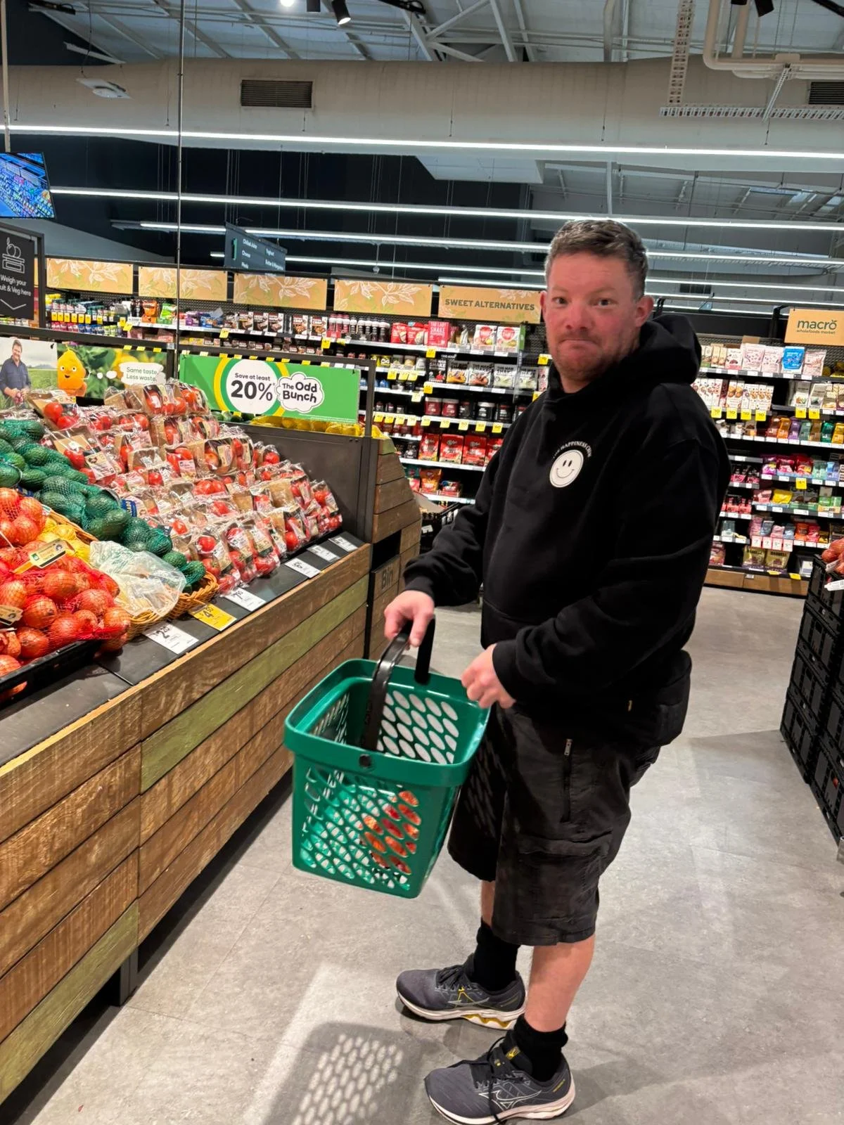 A man shopping for produce in a grocery store, holding a green basket. The produce section has apples and other fruits on display.