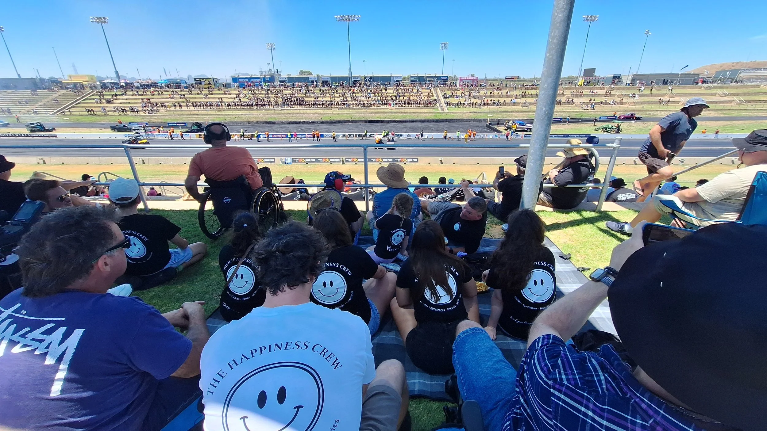 Crowd gathered at drag racing event on sunny day, with spectators seated on grassy area, watching race cars on the track.