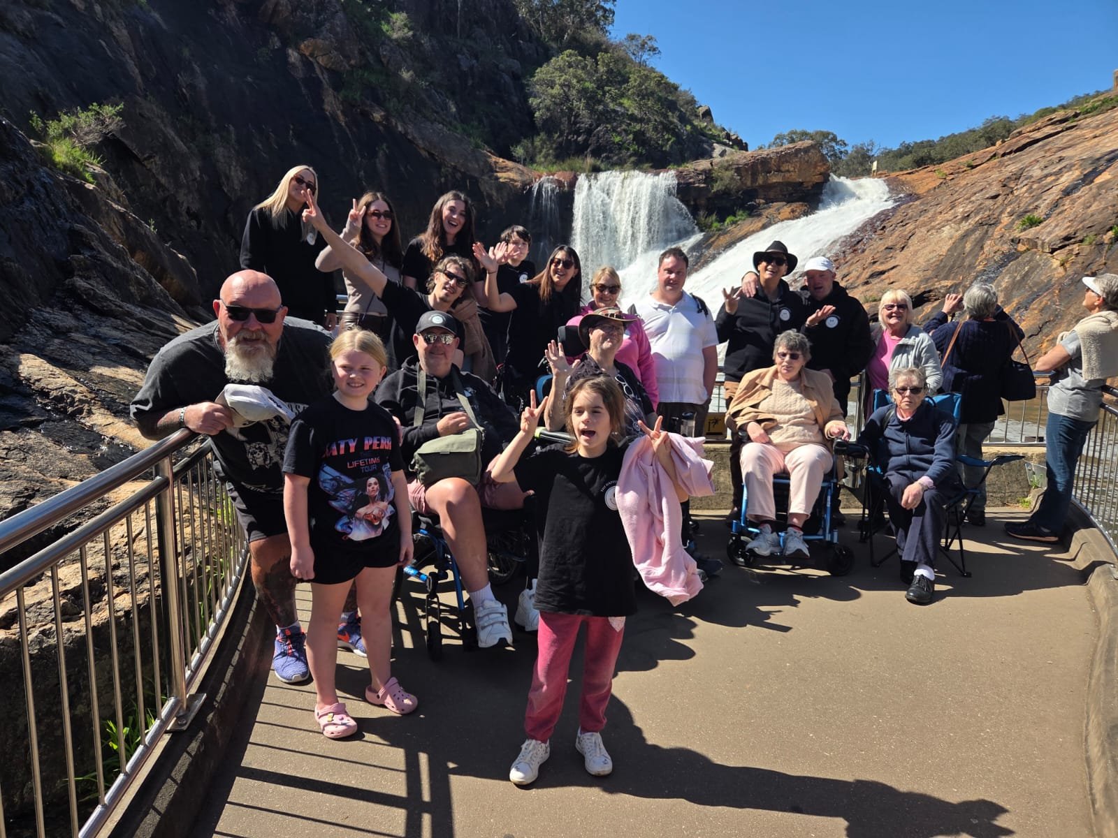 A group of people, including children and elderly, poses for a photo at a waterfall viewing area. The background features a tall waterfall cascading down rocky cliffs under a clear blue sky, with some greenery on the rocks.
