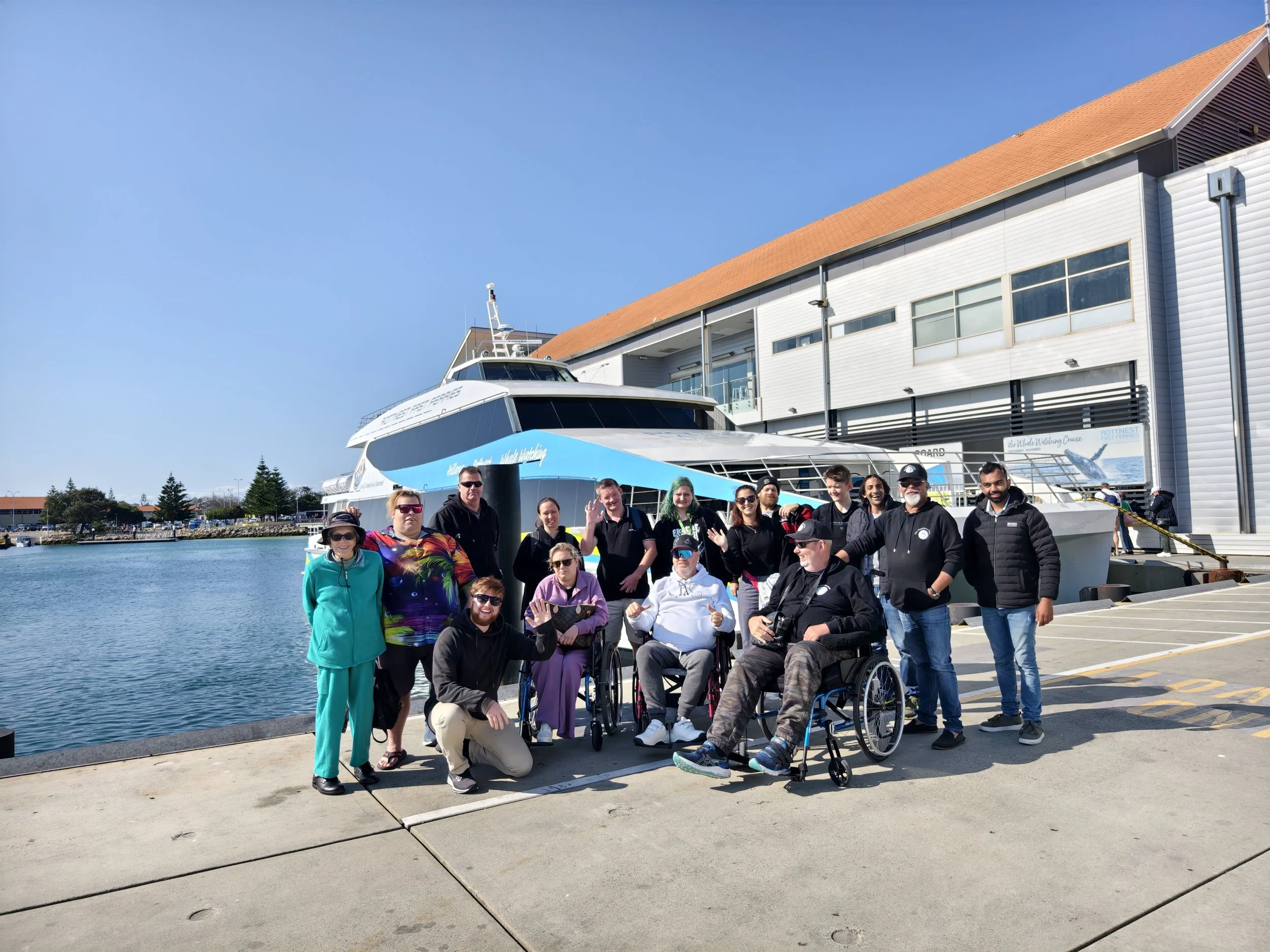 Group of people at the dock in front of a large yacht, sunny day, some individuals in wheelchairs, posing for a photo.