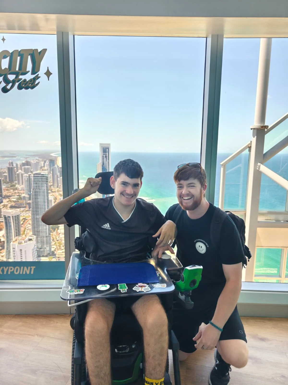 Two men smiling and posing for a photo at a high altitude with a cityscape and ocean view behind them. One man is in a wheelchair with a tray table, and the other is kneeling beside him.