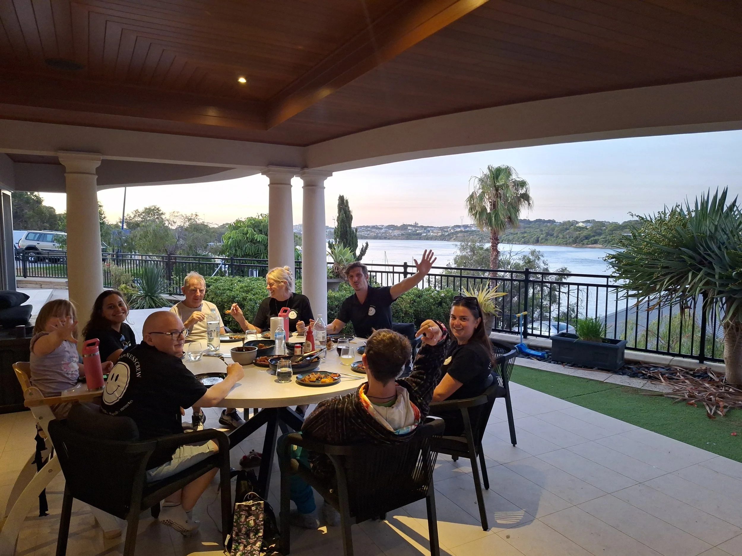 Family and friends gathered around a table on a covered patio, enjoying a meal with a scenic river view in the background.