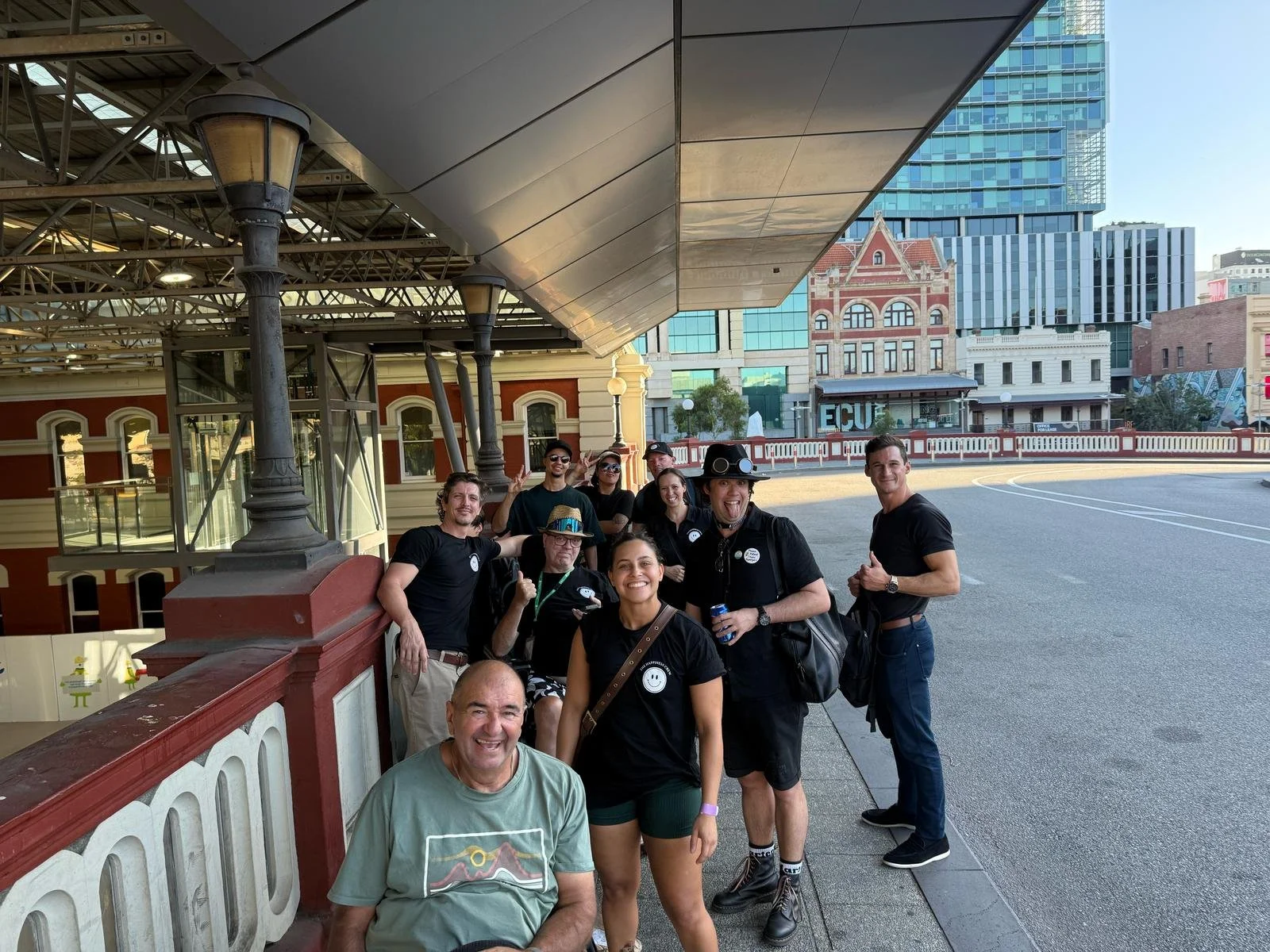 A group of ten people posing together on a city sidewalk appears to be enjoying a sunny day, with some smiling and making various gestures. They are near a building with vintage architecture and modern glass buildings in the background.