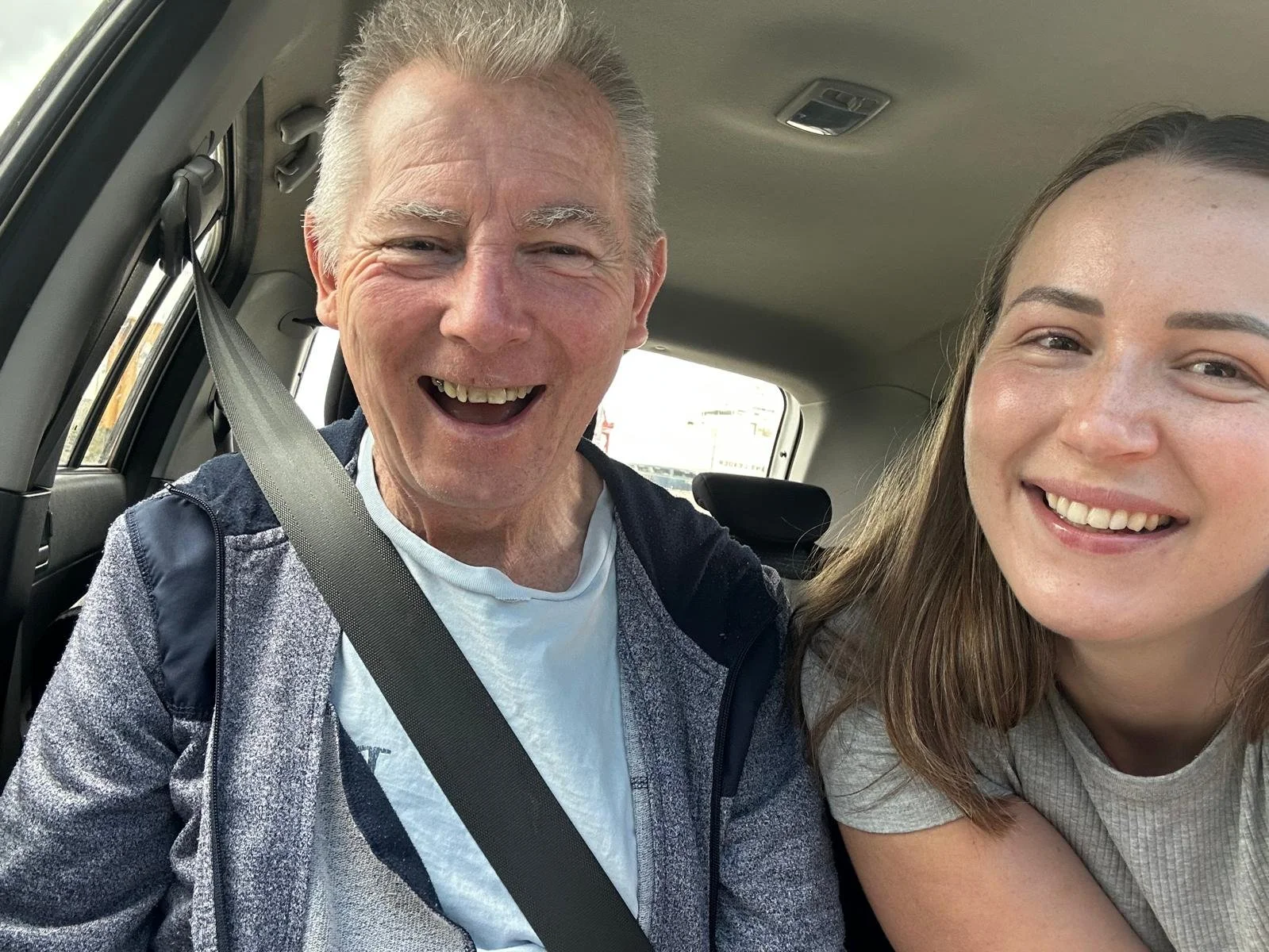 A smiling elderly man and a young woman sitting inside a vehicle, both looking at the camera.