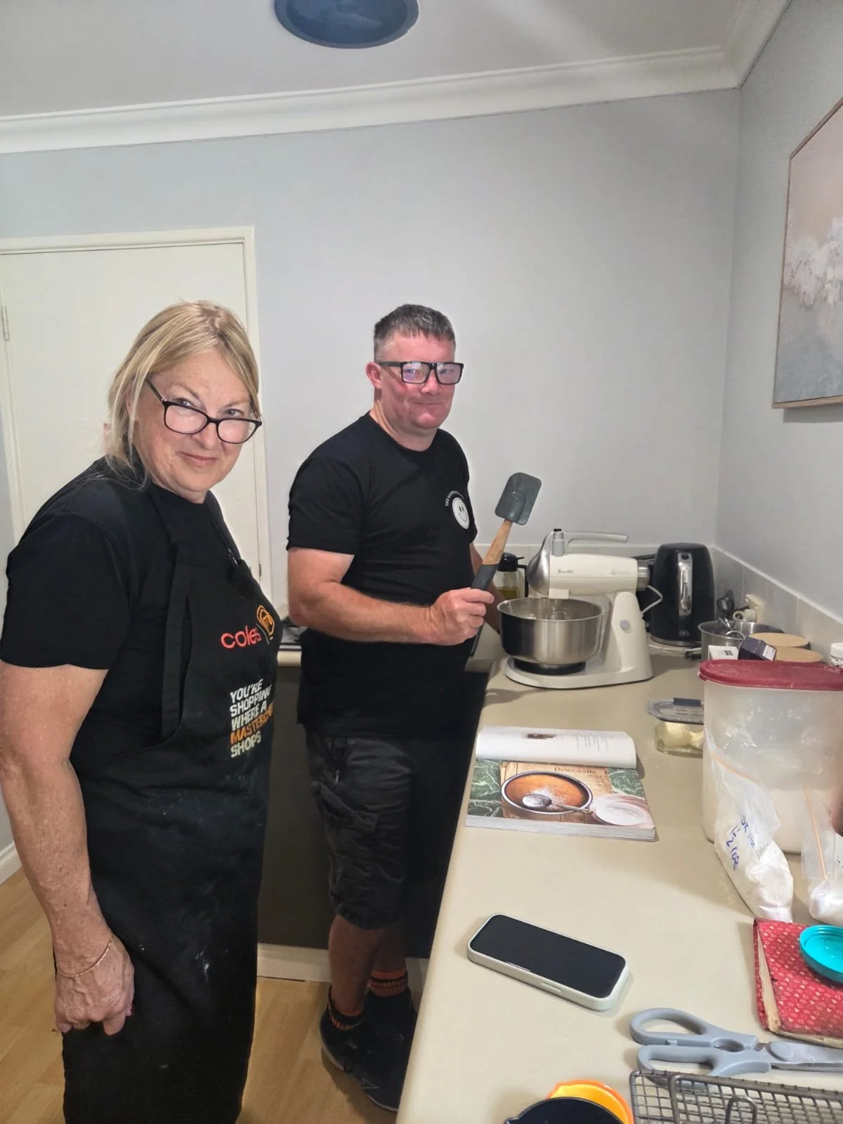 Two people preparing food in a kitchen, one man holding a spatula, and a woman standing next to him. The kitchen counter has a cookbook, a phone, scissors, and various kitchen supplies.