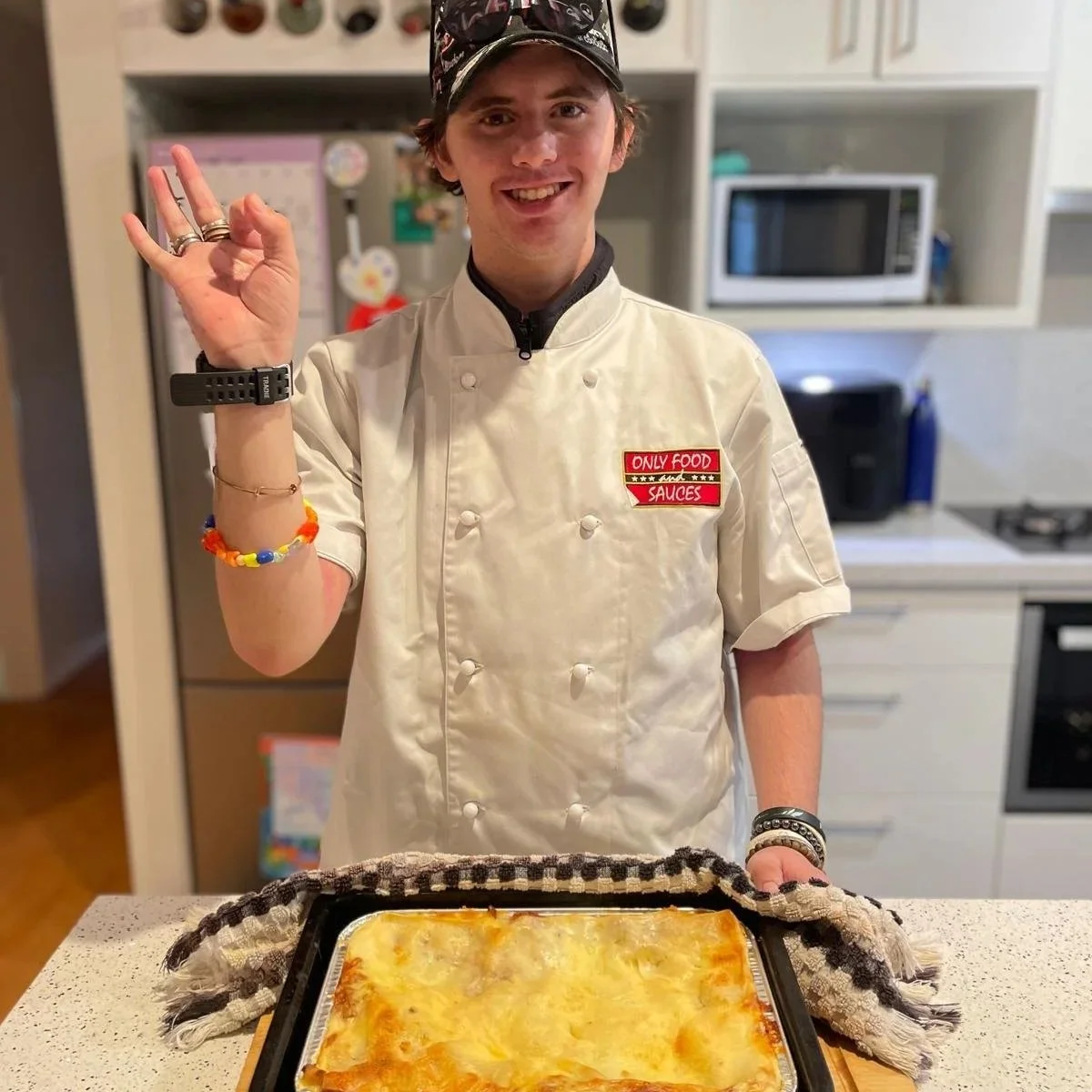 A young man in a chef's coat and cap is smiling and making the 'okay' gesture with his right hand. He is standing in a kitchen behind a tray of freshly baked cheesy lasagna.