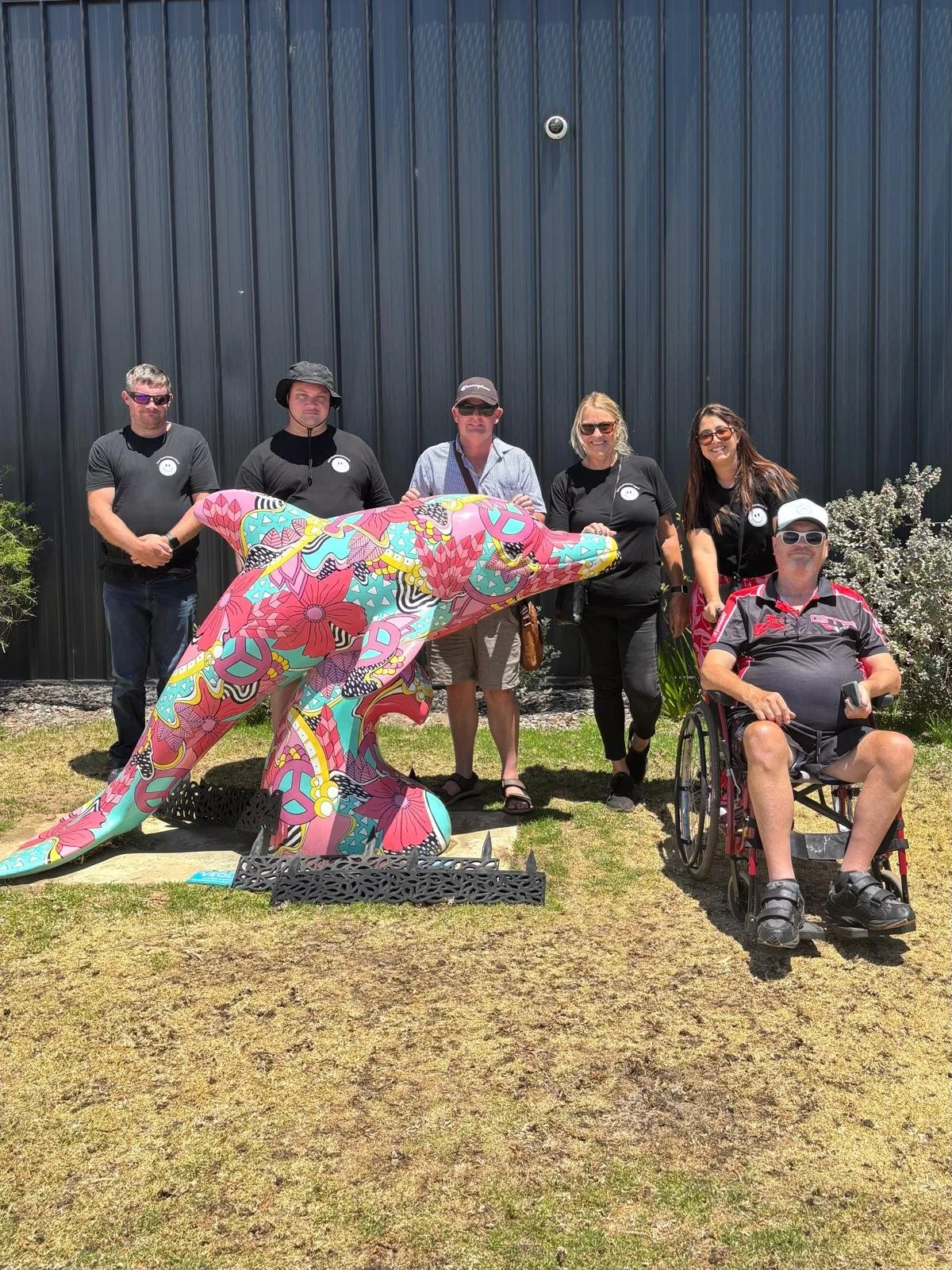 Group of six people standing outdoors, with one man seated in a wheelchair, behind a colorful dolphin sculpture, in front of a dark blue metal building.