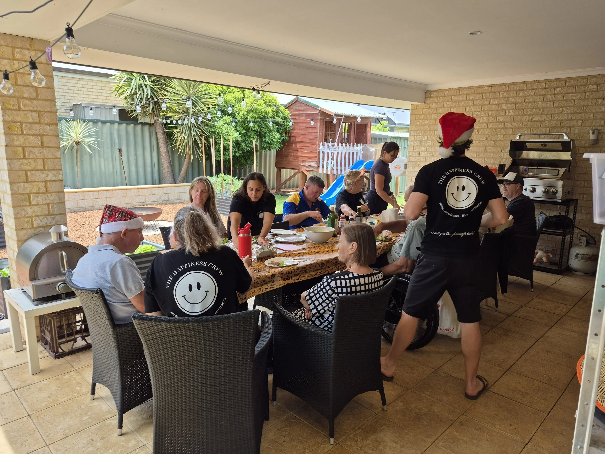 Group of people celebrating Christmas at a backyard gathering with festive hats and holiday decorations, including a Santa hat and Christmas-themed table.
