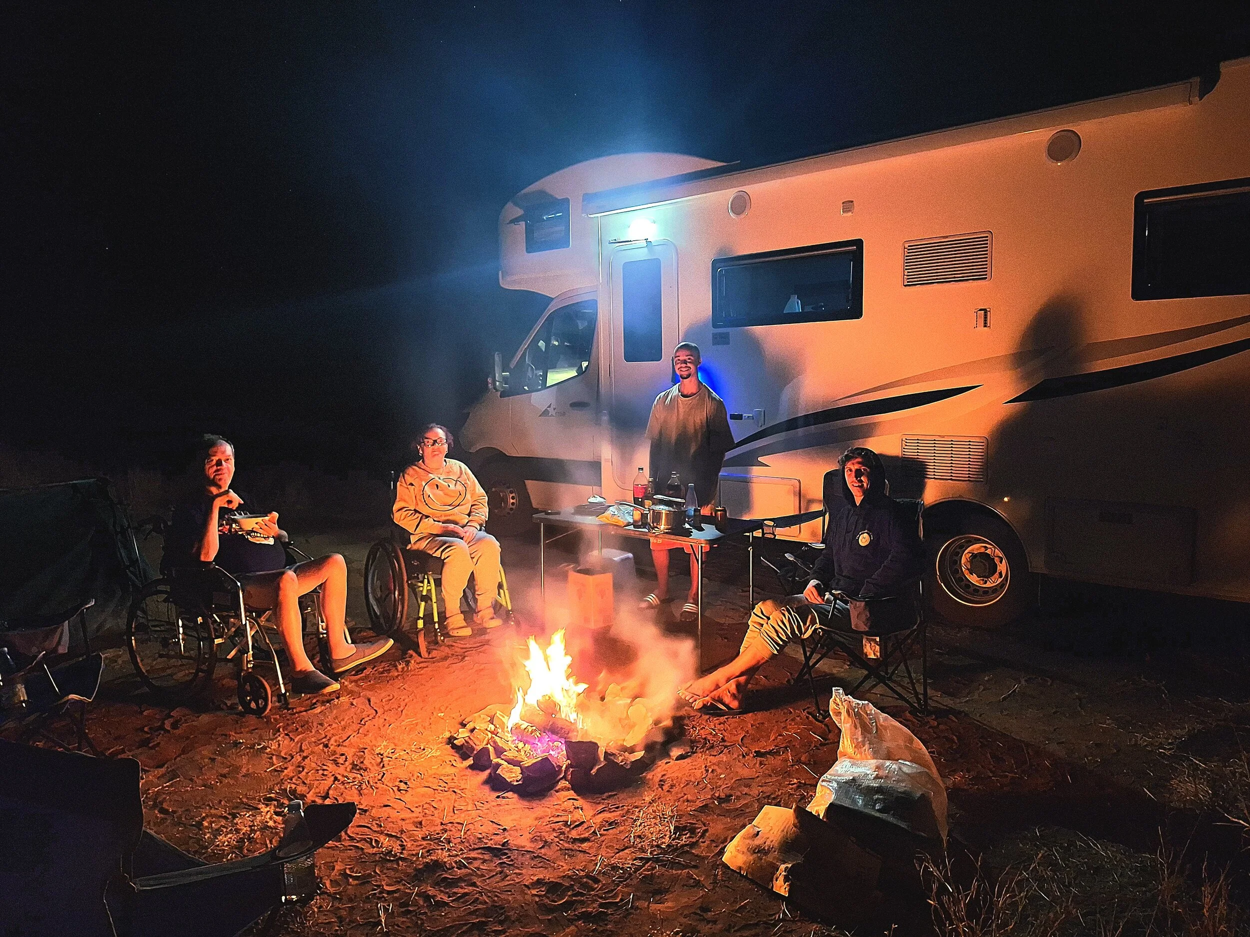 Four friends sitting around a campfire at night with a large RV in the background. Two are in wheelchairs, one is sitting on a camping chair with feet near the fire, and another is standing behind the table with drinks on it. The scene is illuminated by the fire and RV lights.