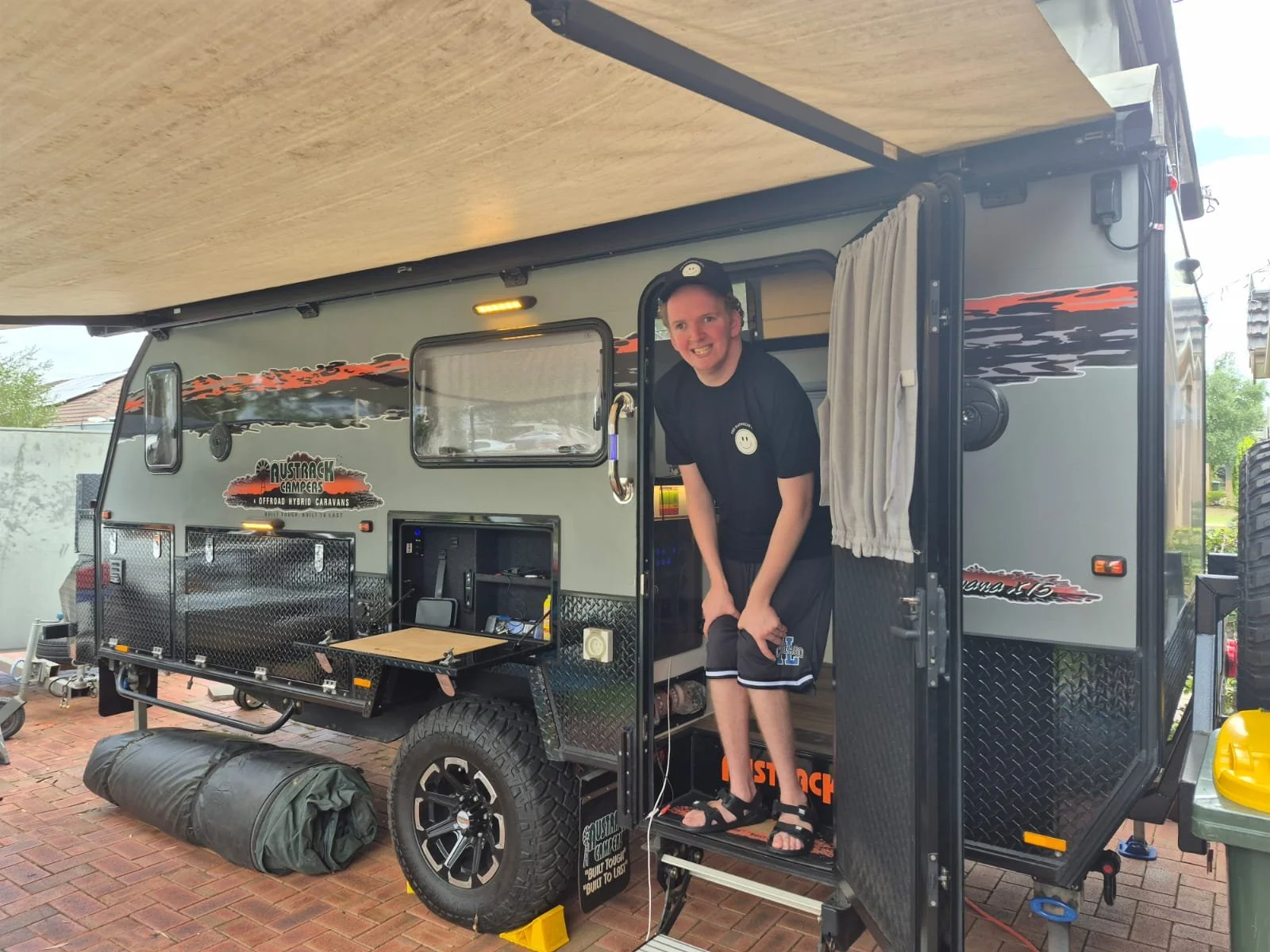 A young man smiling and leaning out of a camper trailer doorway. The camper has a gray exterior with orange and black decals, and is parked on a brick driveway. There is a rolled-up sleeping bag or tent on the ground in front of it.