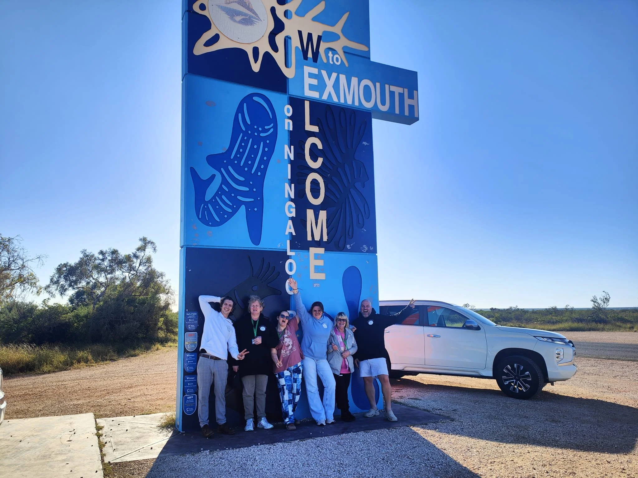 Group of six people standing in front of a large sign for the town of Exmouth, featuring a fish and a seashell, with a white vehicle in the background, under a clear blue sky.