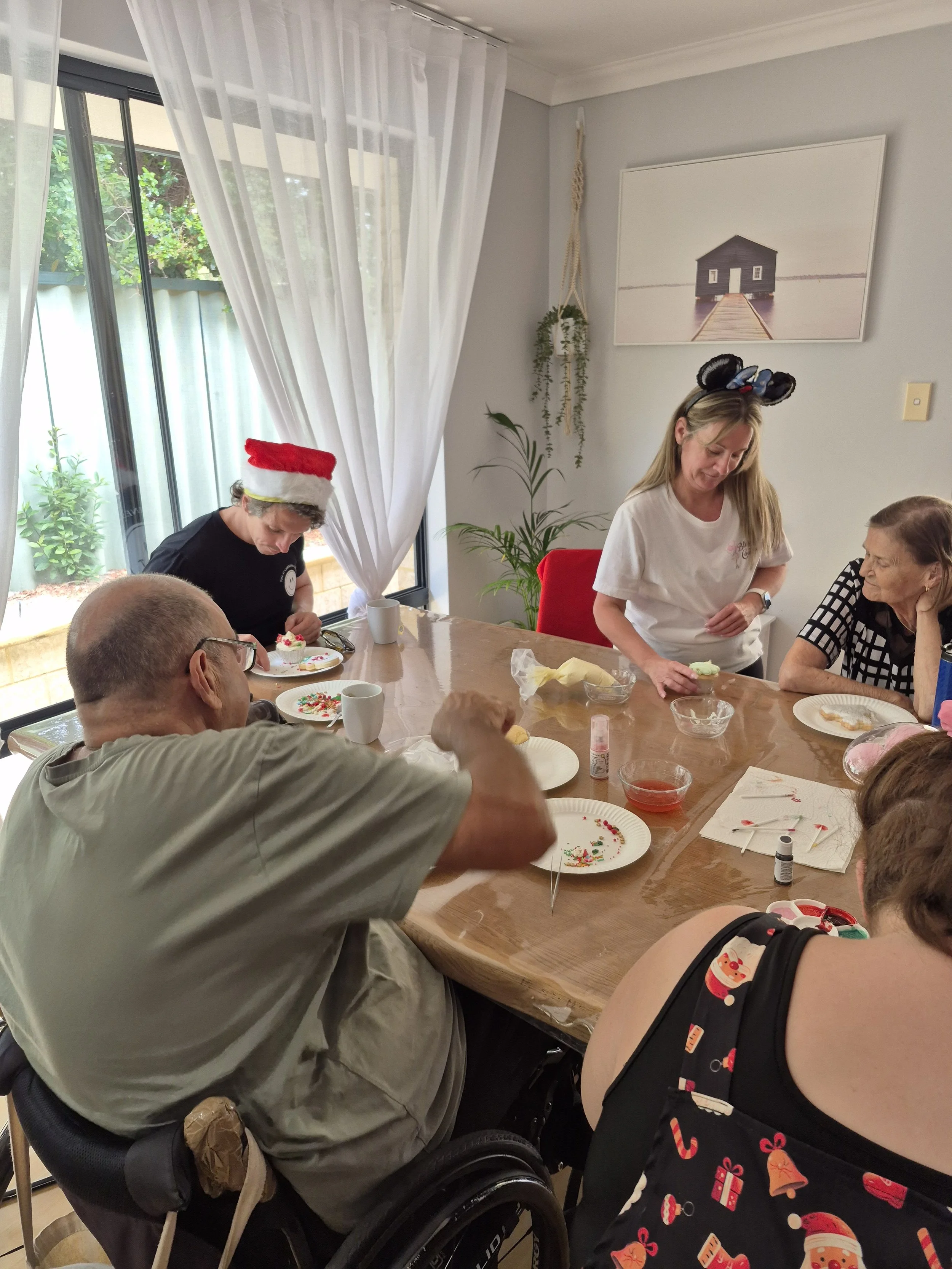 People decorating cookies at a holiday gathering around a wooden table, with some wearing Christmas-themed accessories like a Santa hat and reindeer ears, in a bright room with natural light from large windows and white curtains.