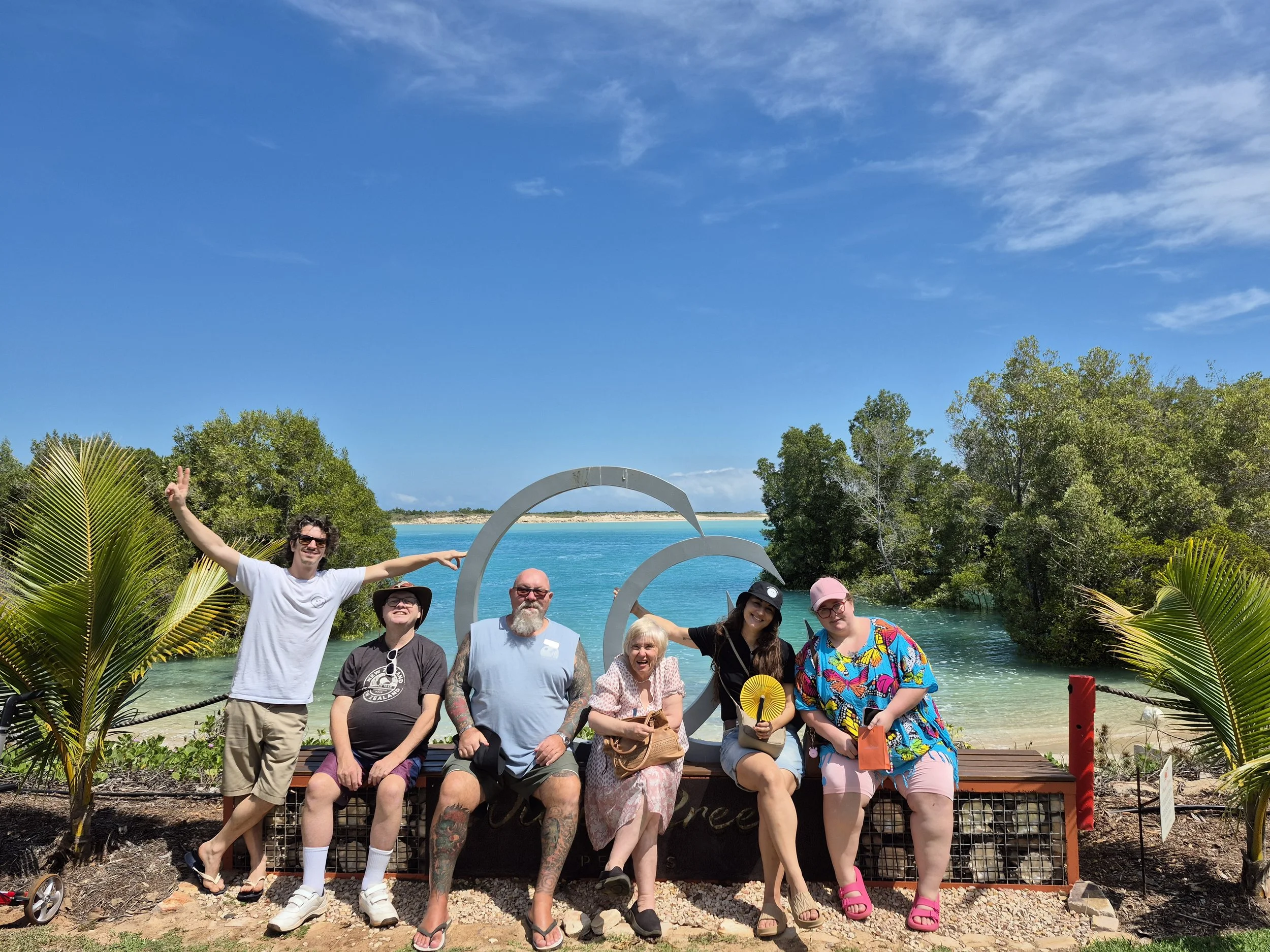 Group of seven people sitting and standing in front of a scenic beach background with clear blue water, green trees, and a bright sky, posing for a photo with some making gestures and holding accessories.