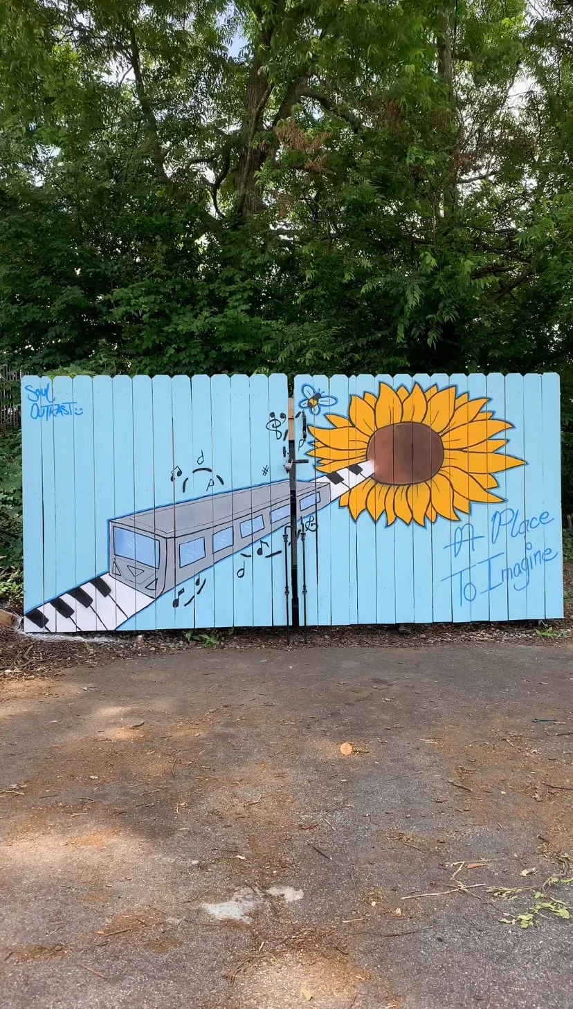 A street art mural painted on a light blue wooden fence features a train coming out of a sunflower, with musical notes and a bee near the flower. The mural includes the phrases 'Soul Outcast!,' 'A Place to Imagine,' and some musical symbols.