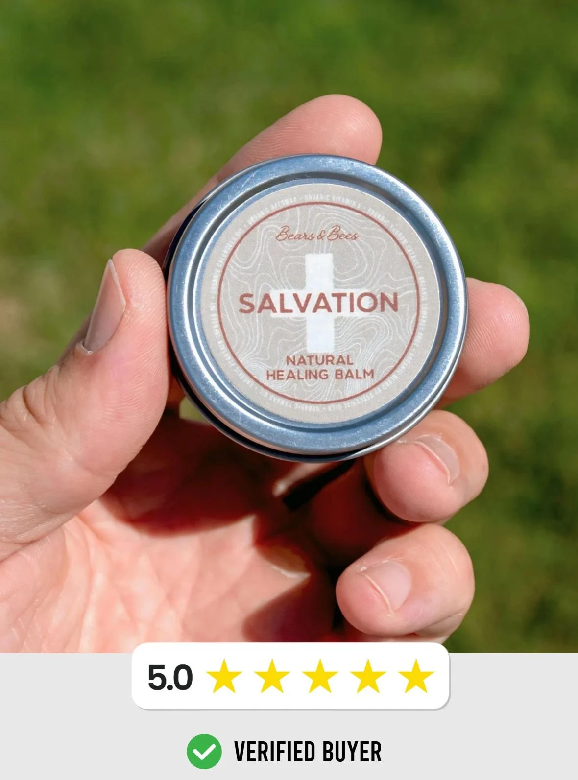 A hand holding a small round tin of Alpine Ridge Soap Company's Salvation natural healing balm against a grassy background.