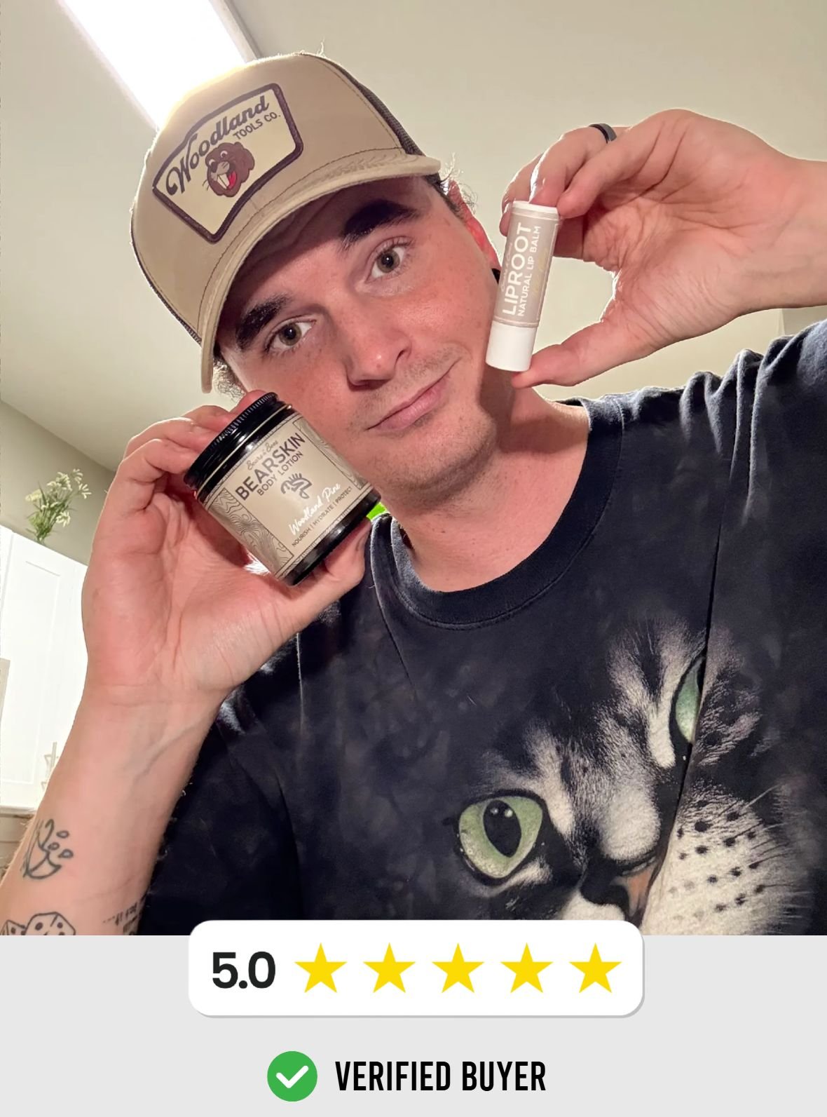 Young man with a tattoo on his arm, wearing a black T-shirt with a cat face graphic, holding a jar of skincare product and a lip balm tube, smiling at the camera, inside a home.