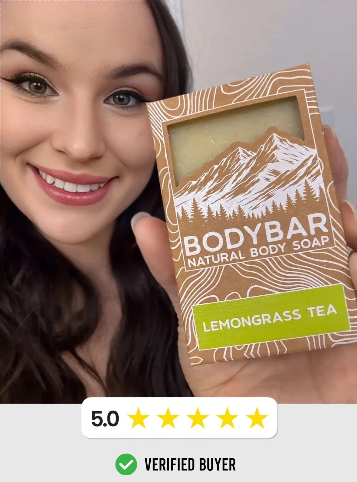 A woman with long dark hair and makeup smiling while holding a bar of natural body soap with mountain and forest illustrations on the packaging, labeled 'Lemongrass Tea'.