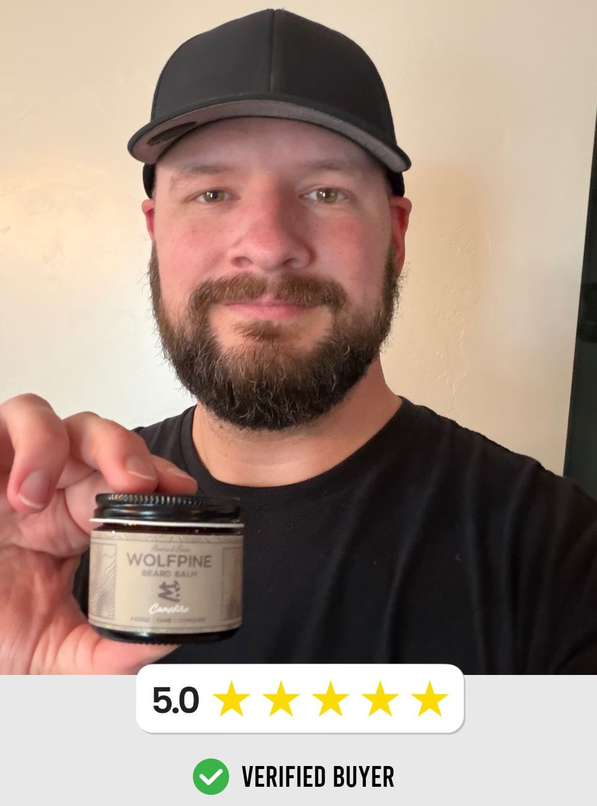 A man with a beard and mustache wearing a black cap and black shirt holding a jar of Wolfpine beard balm, smiling at the camera.