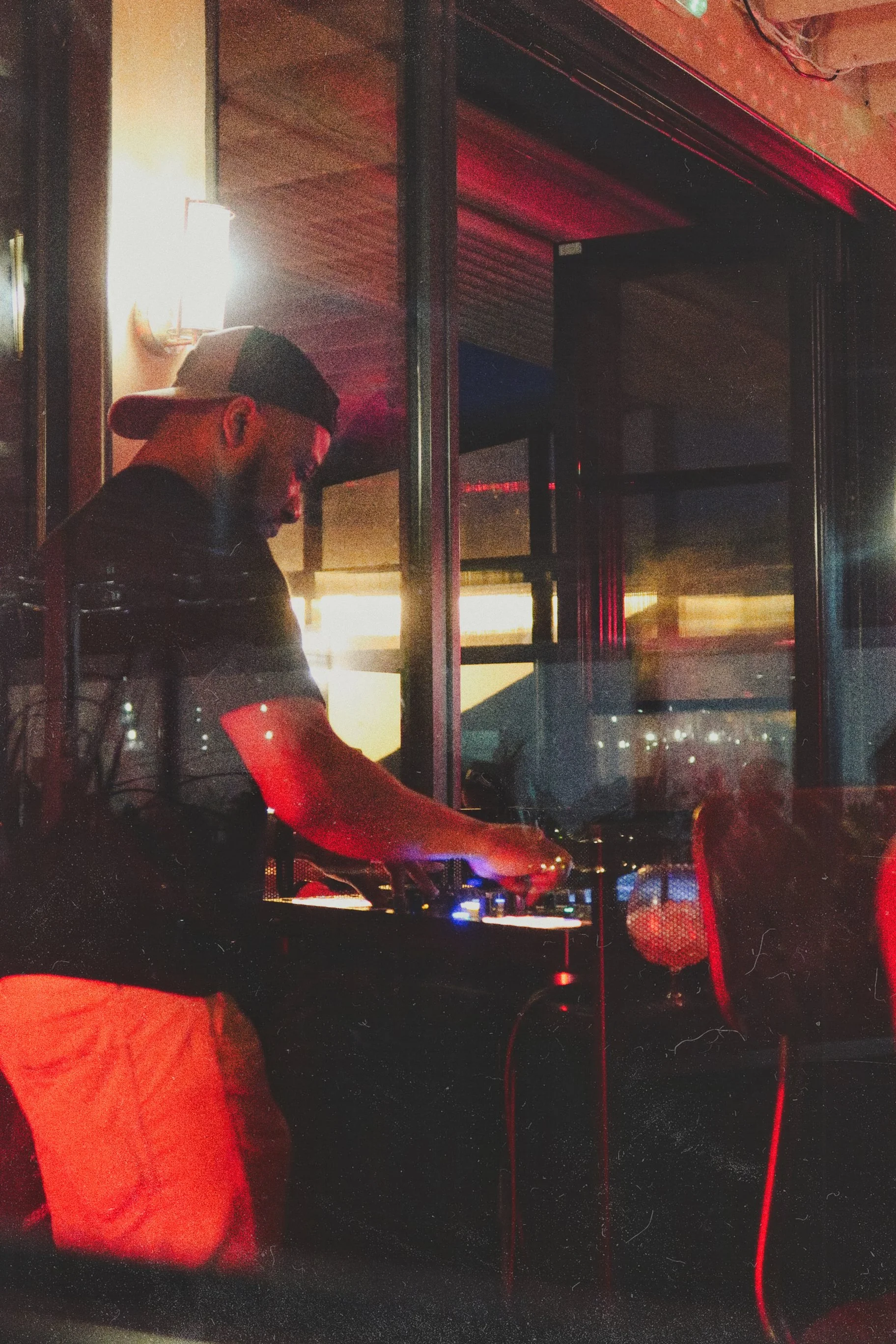 DJ wearing a cap and black shirt, mixing music on turntables in a dimly lit indoor venue with red lighting and reflective surfaces.