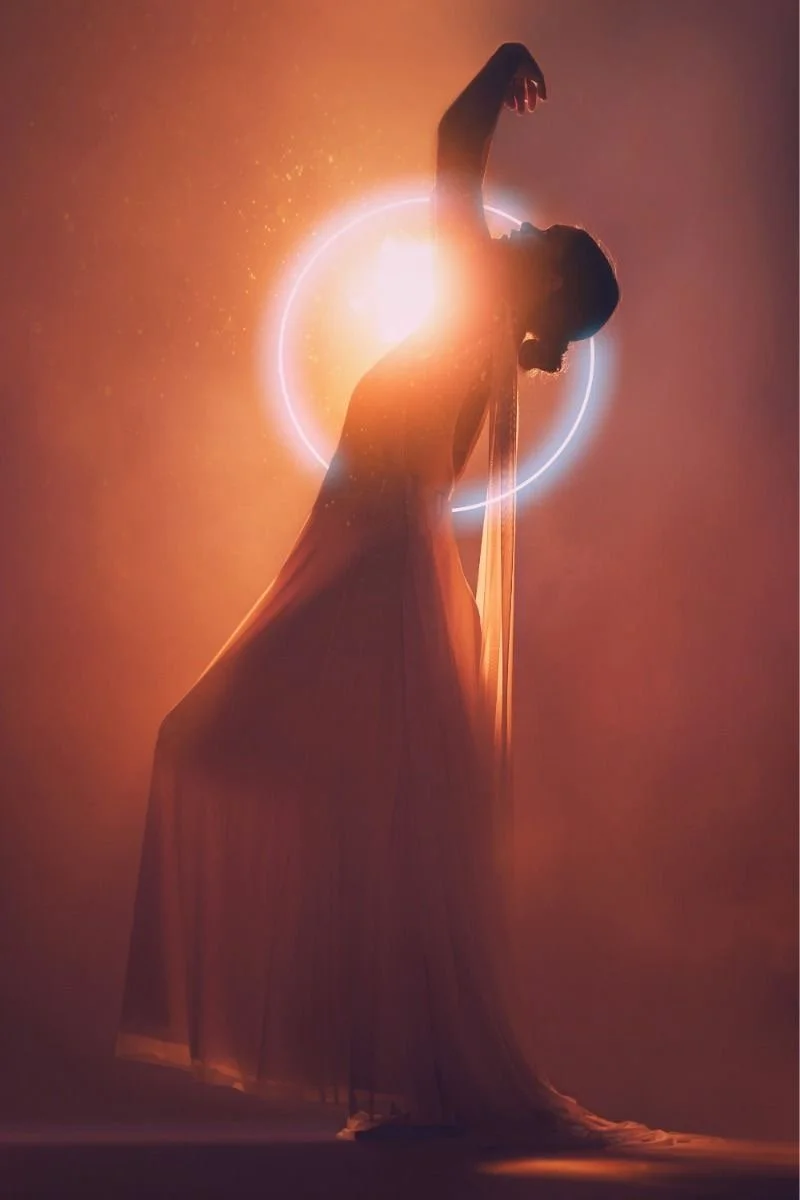 A silhouette of a woman in a flowing dress, performing a dance pose against a cosmic background with a bright light and rings behind her.