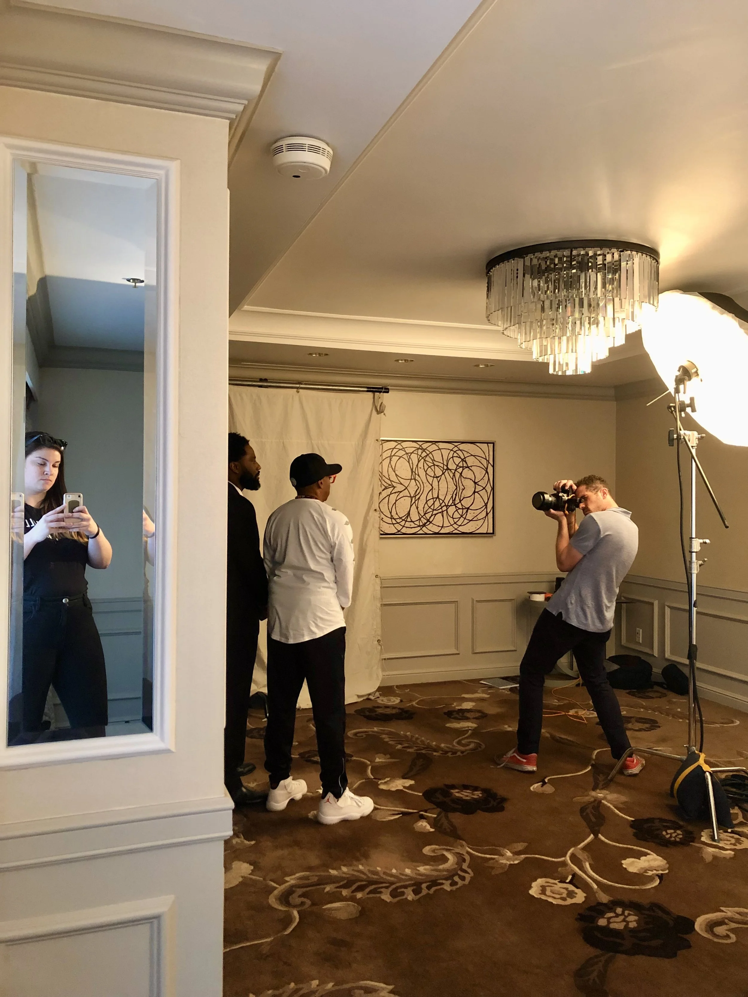 People taking photos in a professional photo shoot setup with a photographer, a woman taking a picture on her phone, and three individuals posing in front of a white backdrop, indoor decorated room with chandelier, abstract wall art, and plush carpet.