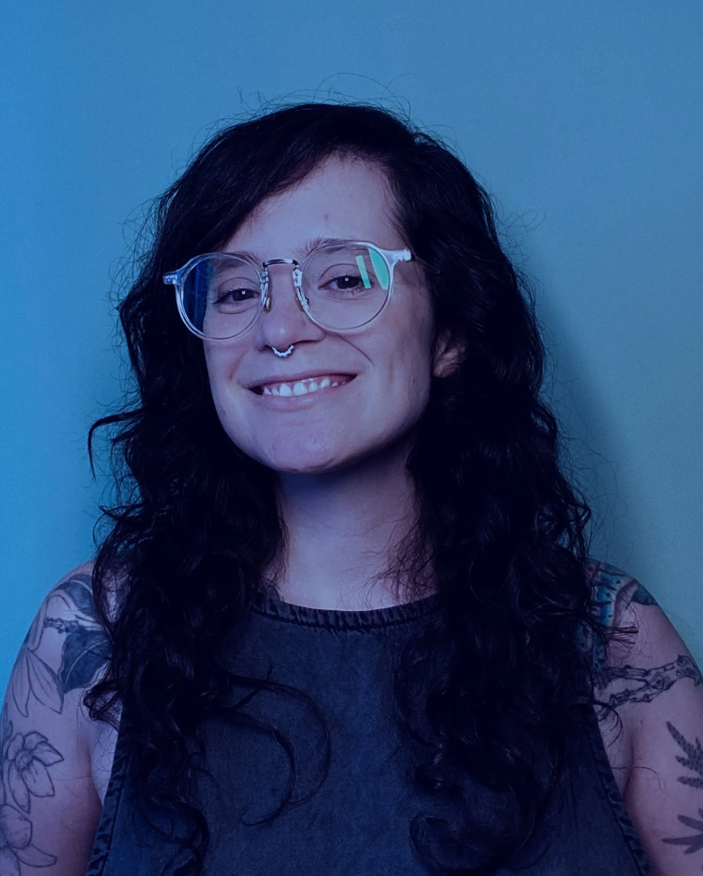 Retrato de uma mulher com cabelo cacheado escuro sorrindo, usando óculos transparentes, com uma blusa preta sem mangas e tatuagens que aparecem nos dois braços, fundo azul claro.