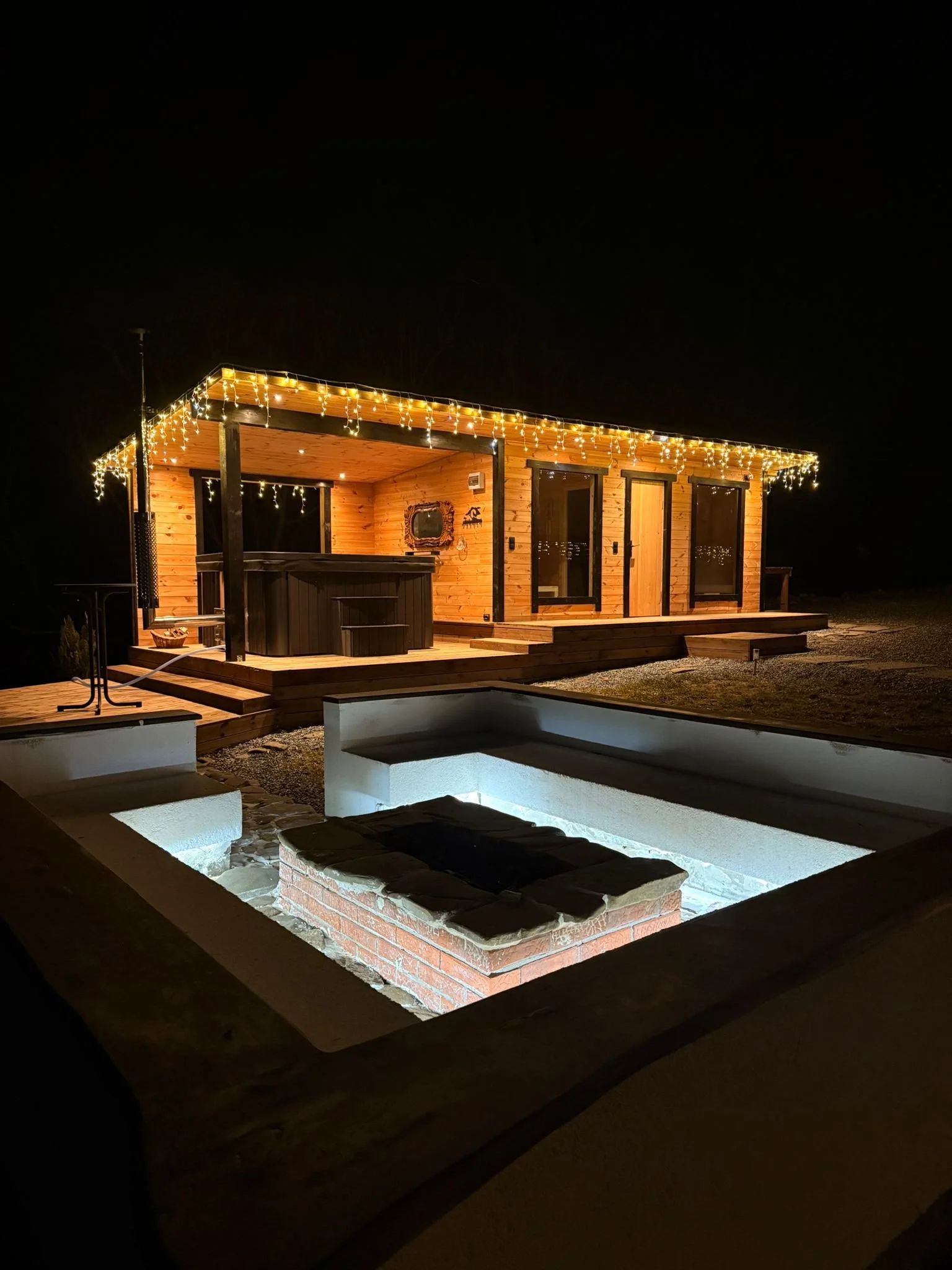 Wooden house or cabin at night decorated with string lights, with a hot tub on the porch and a small pool or fire pit in the foreground.