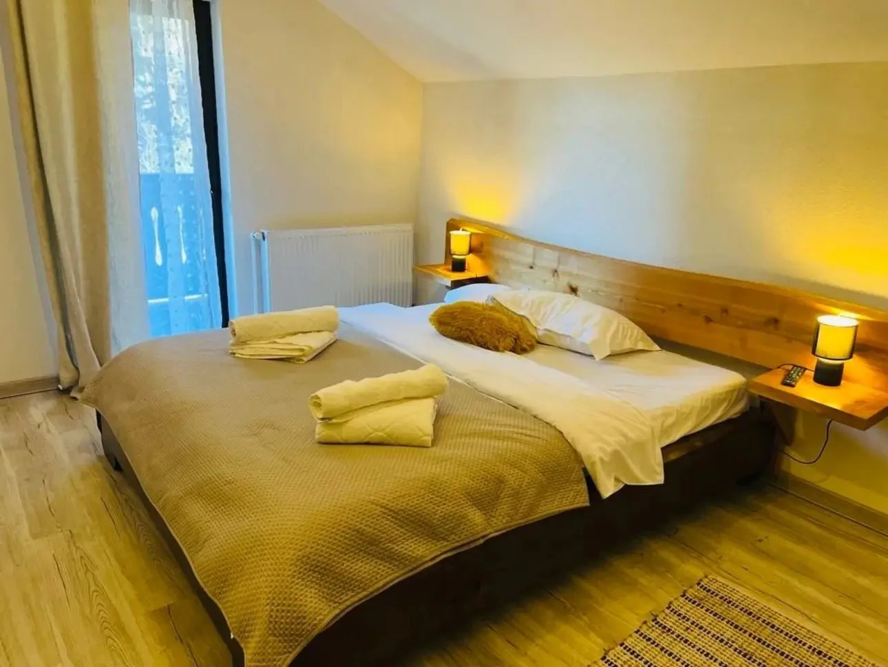 A cozy bedroom with a large bed, two bedside tables with lamps, a wooden headboard, and a sliding glass door with curtains leading to a balcony.