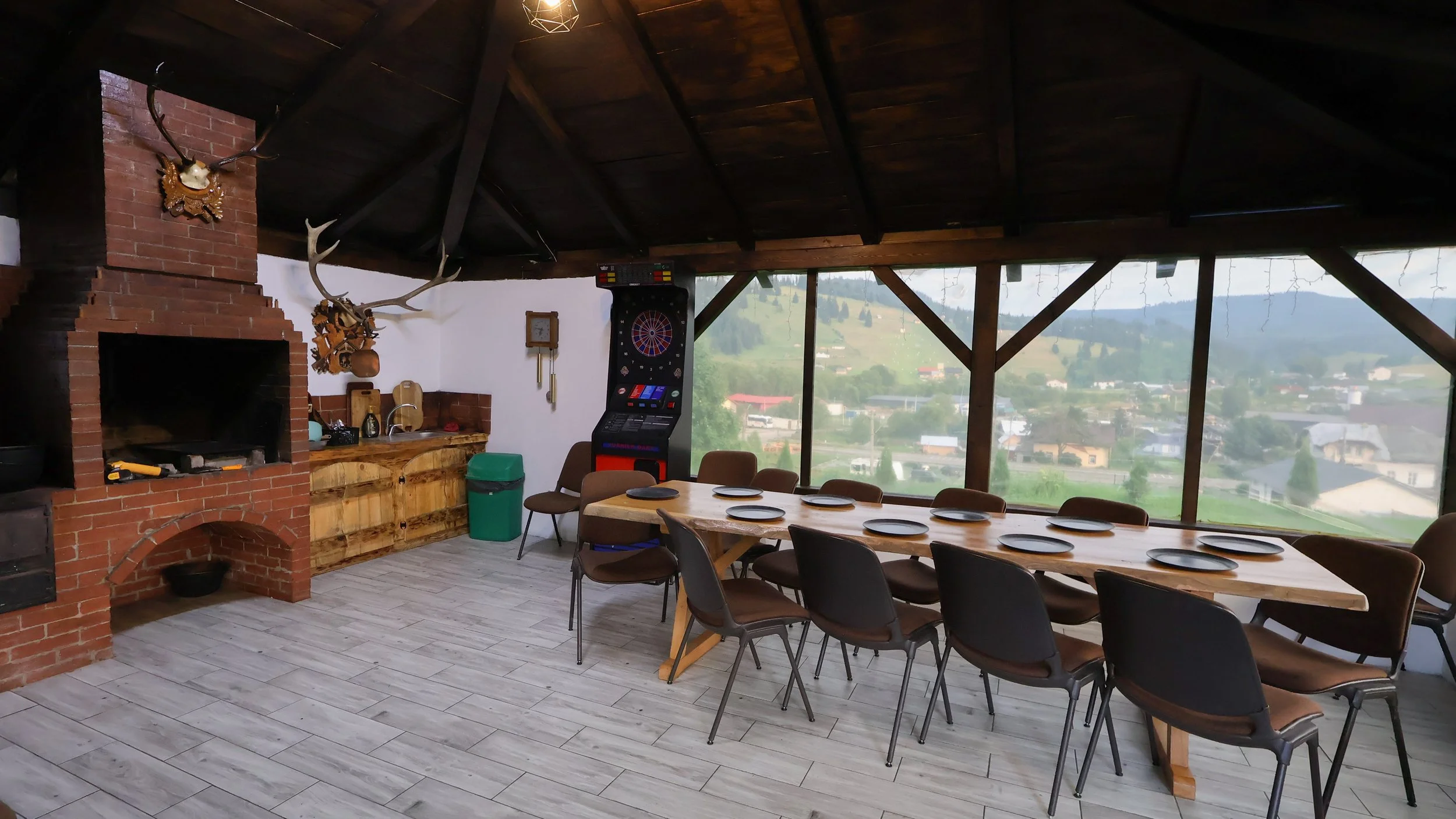 Indoor dining area with a long wooden table set with plates, surrounded by chairs. There is a brick fireplace with a mounted deer head and antlers, a dartboard, a wooden countertop with kitchen utensils, a green trash bin, and large windows showing a scenic view of a rural landscape with houses and hills.