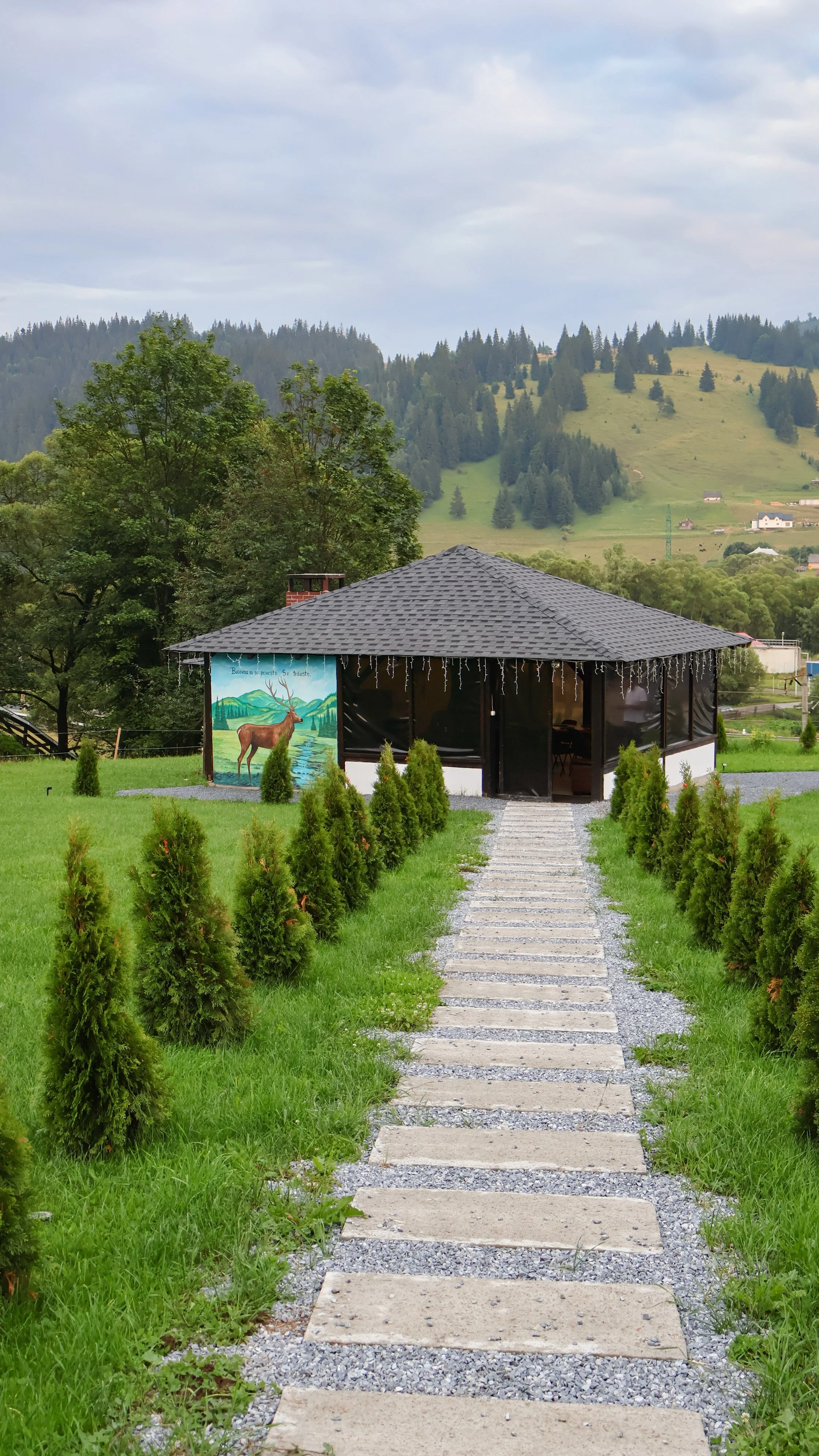 A pathway of rectangular concrete and gravel stepping stones leads to a small building with black screened sides and a gray shingled roof. The building has holiday string lights hanging from the roof edges and a mural of a deer painted on the side. Small bushes line the pathway. In the background, there are green hills with scattered trees and houses, and a cloudy sky.