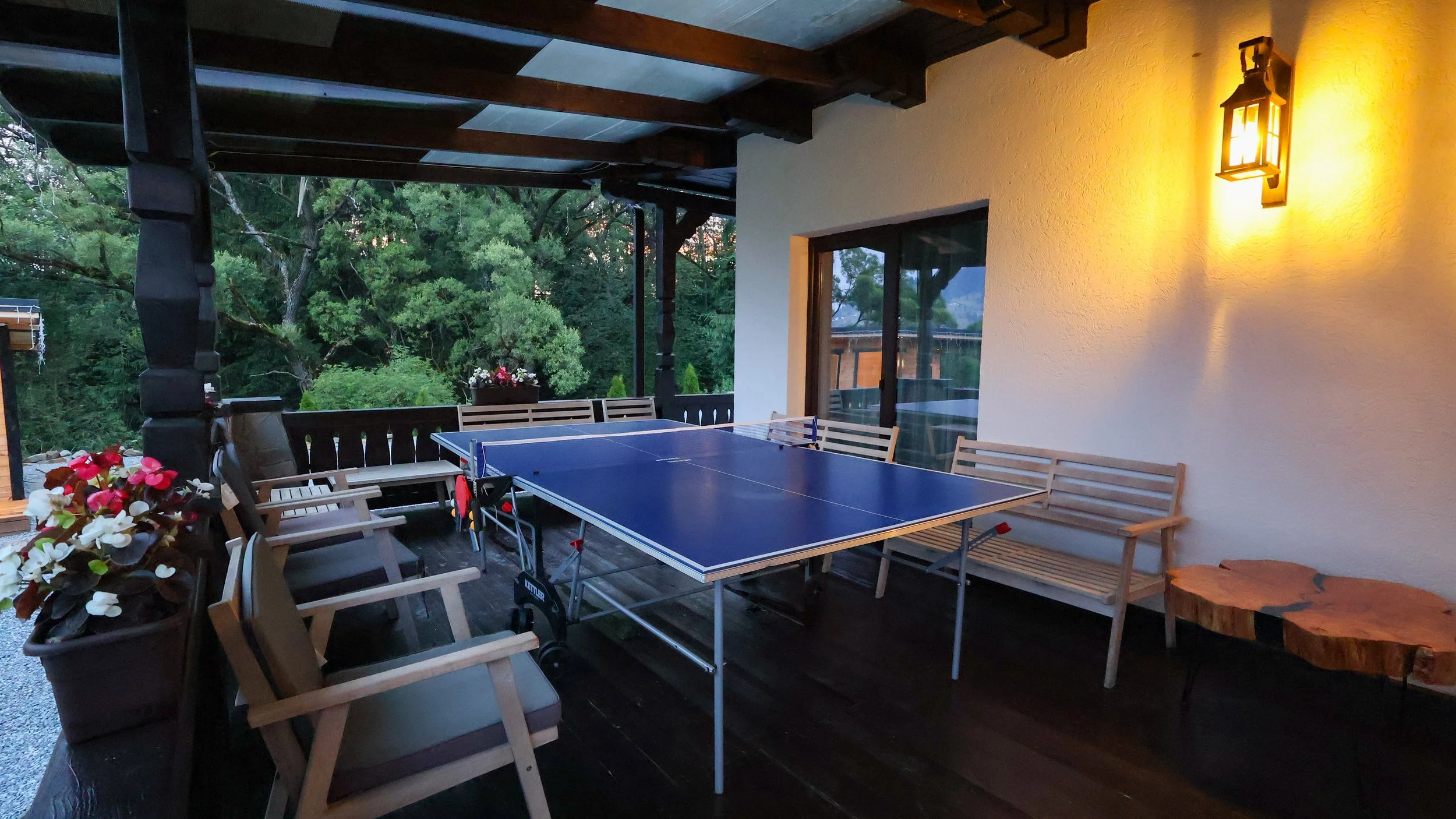 An outdoor porch with a dark wood ceiling and beams, beige stucco wall, and a wall-mounted lantern light. There is a blue ping pong table with wooden benches and chairs around it. The porch overlooks green trees and has potted flowers.