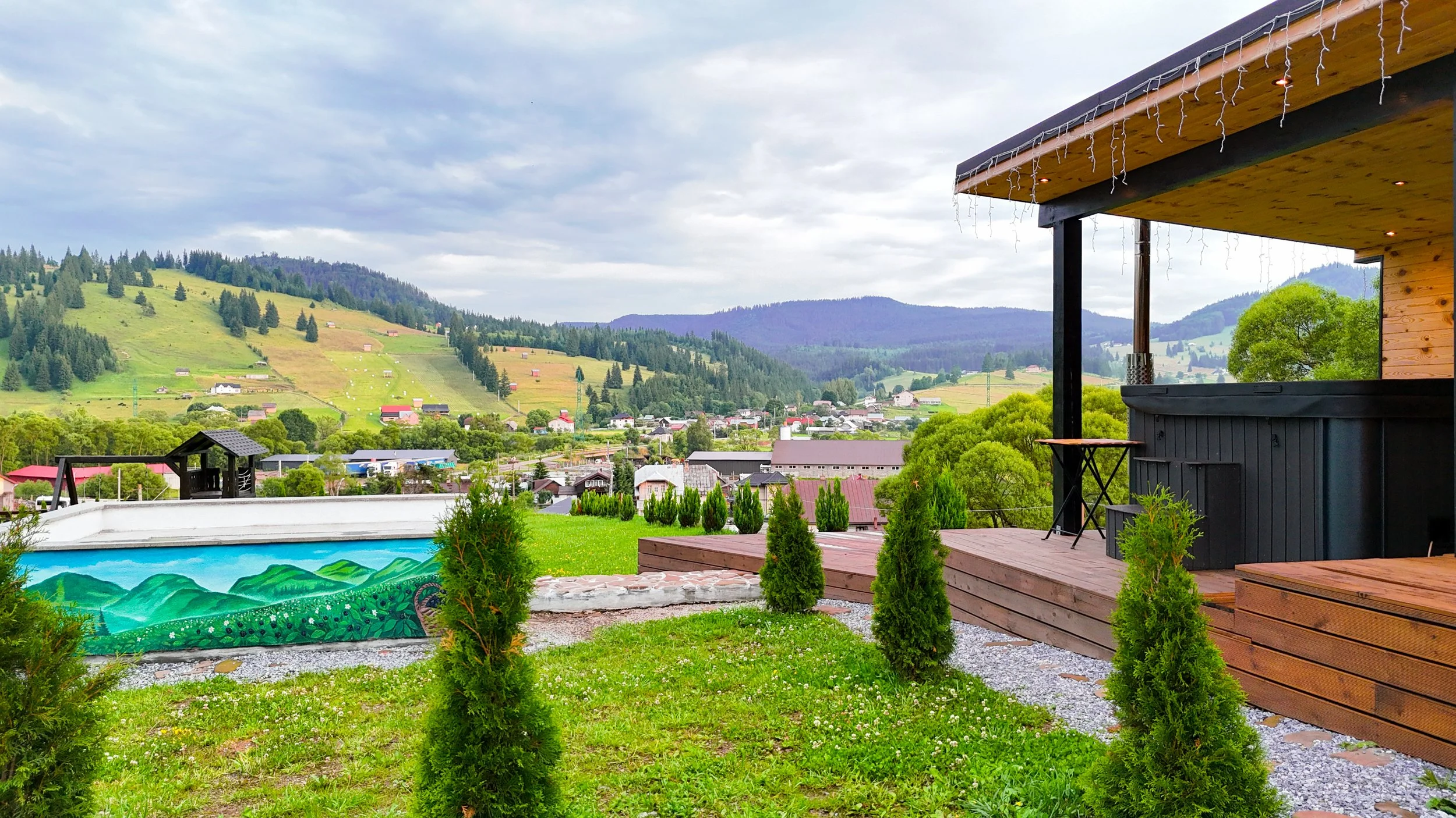 A view from a deck showing a grassy backyard with small trees, overlooking a town with houses, rolling green hills, and mountains under a cloudy sky.