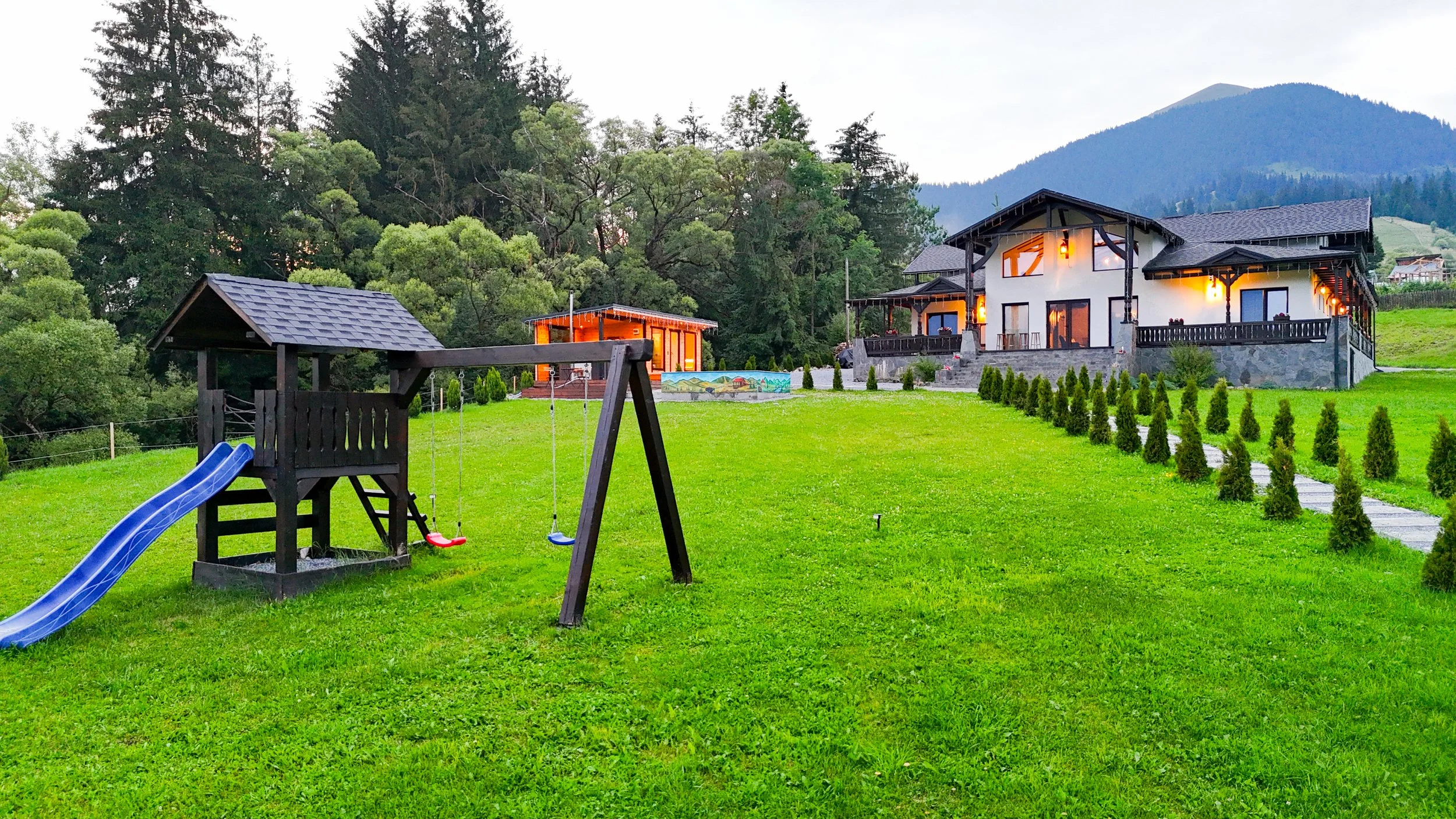 A spacious backyard lawn with a blue slide and swing set, a small wooden shed with a shingled roof, and a large two-story house with outdoor lighting, set against a backdrop of green trees and mountains.