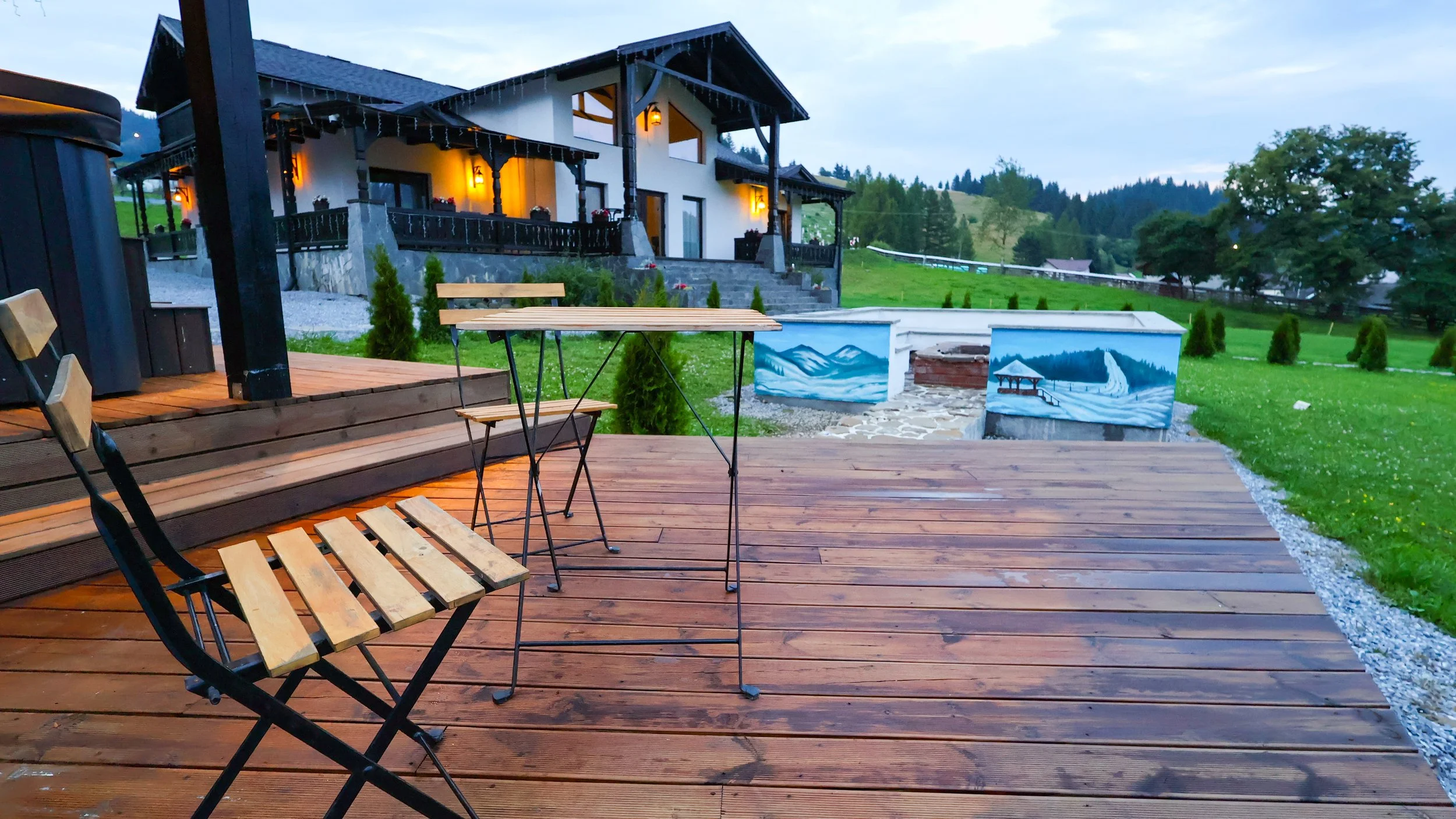 A wooden deck with patio table and chairs in front of a house with lights, set in a green, hilly landscape with trees, and murals painted on structures nearby.