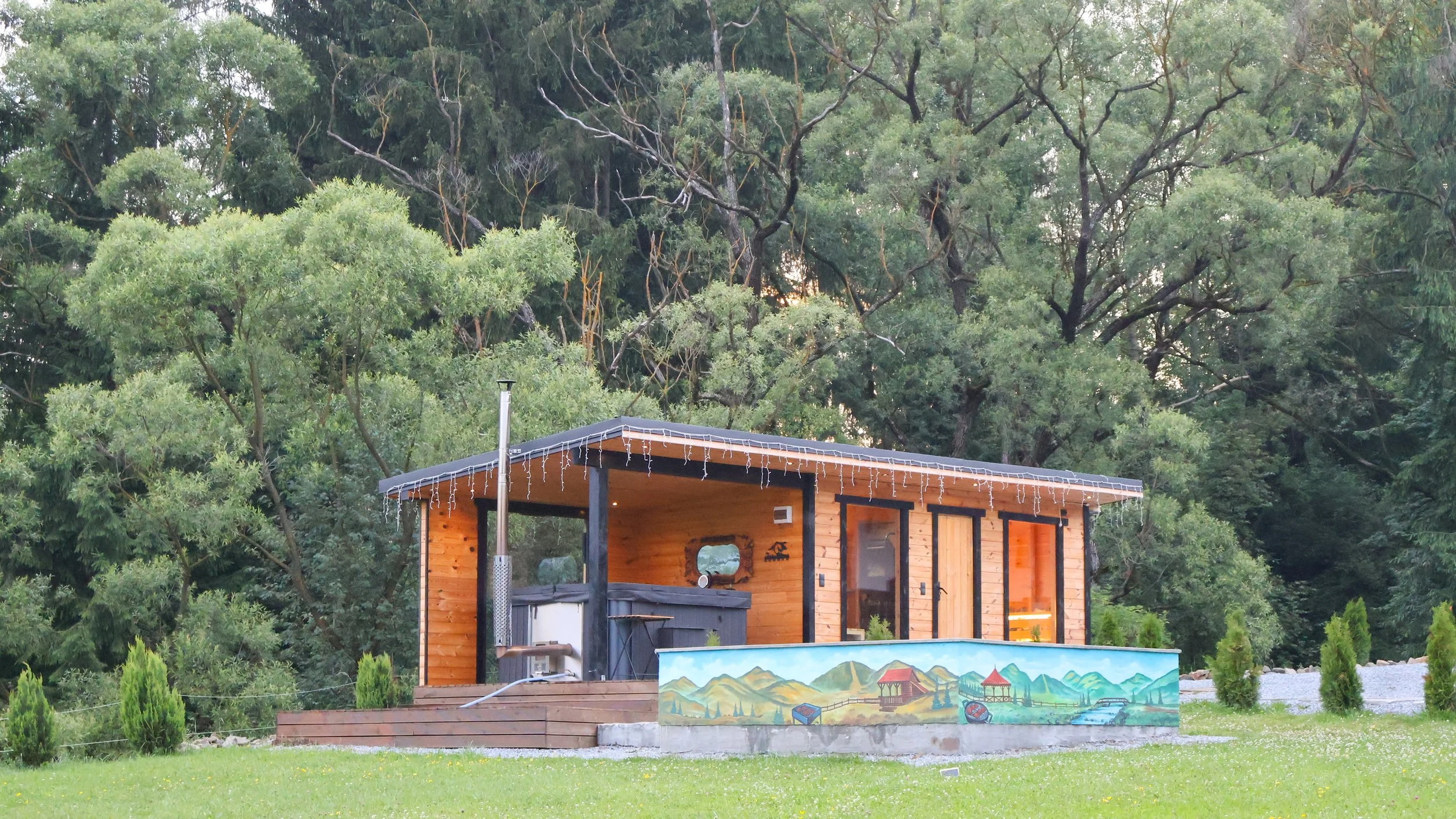 A small wooden cabin with string lights on the roof set in a lush, green area with many trees in the background.