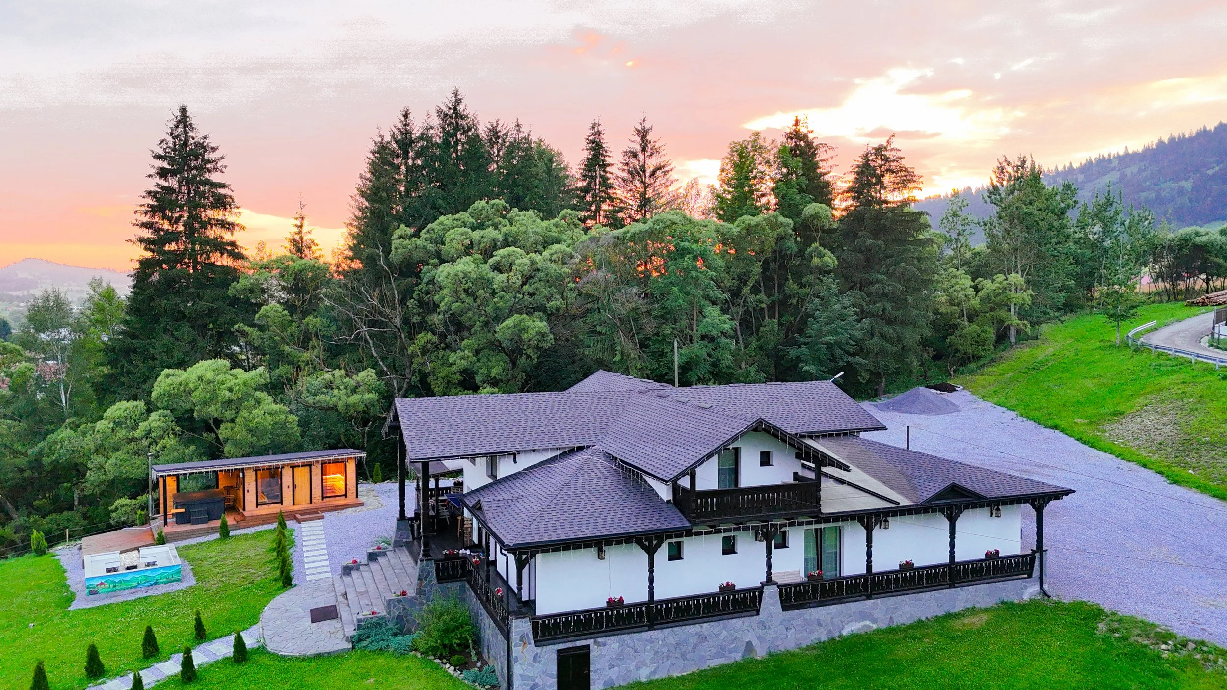 A residential house with multiple covered patios, situated among lush green trees with a mountain and sunset in the background.