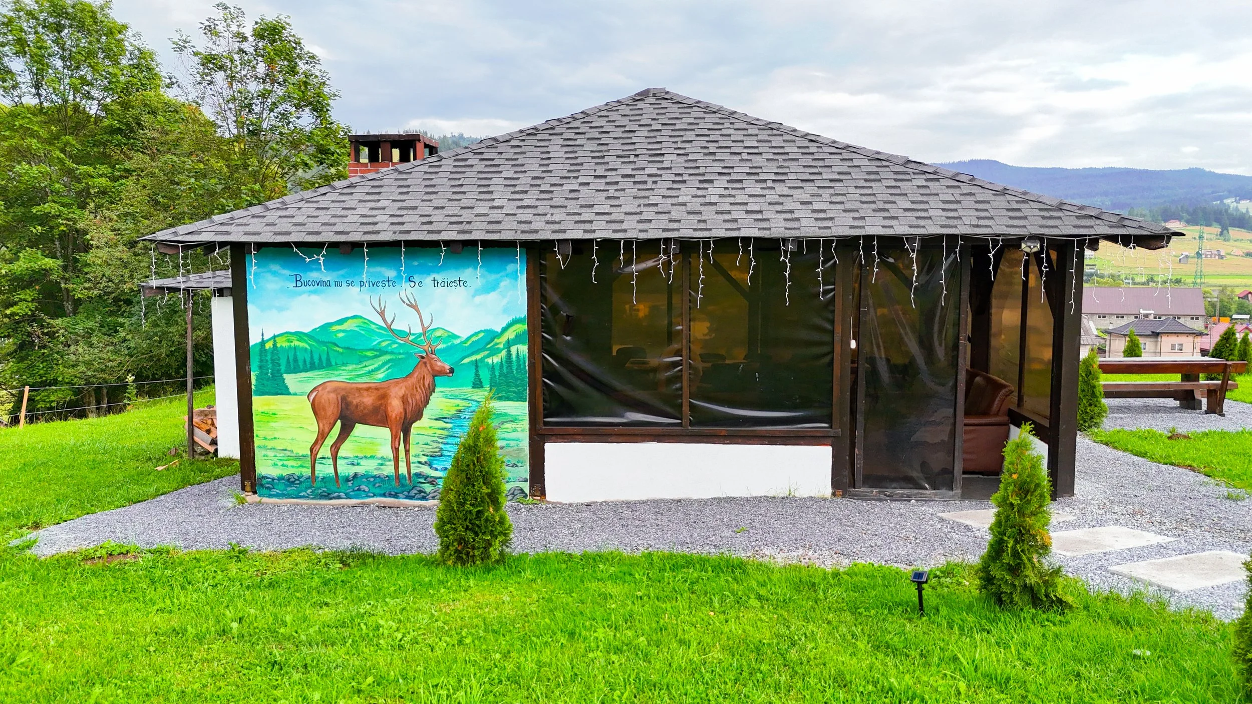 A small outdoor shelter with a gray shingle roof and open sides, decorated with string lights. The front wall has a colorful mural of a brown elk with large antlers, a river, green hills, and mountains, along with the text in Romanian. The setting includes green grass, small bushes, and a gravel pathway, with a scenic landscape of trees, rolling hills, and houses in the background.