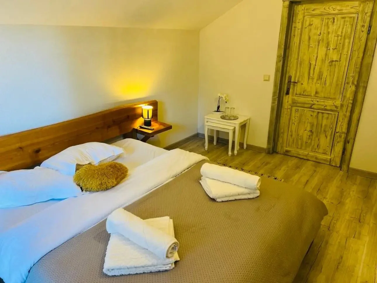 A cozy bedroom with a neatly made bed featuring white pillows and a beige blanket. A small heart-shaped pillow is on the bed. Beside the bed, a wooden nightstand with a black and yellow lamp. In the corner, a white side table with a flower vase and a tray. The room has wooden flooring and a large wooden door.
