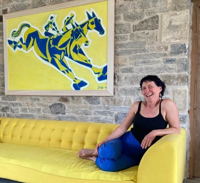 Maria Mountain - Intimacy and Connection Coach. sitting on a yellow couch, smiling, in front of a stone wall with a colourful painting of three horses being ridden by jockeys hanging above.