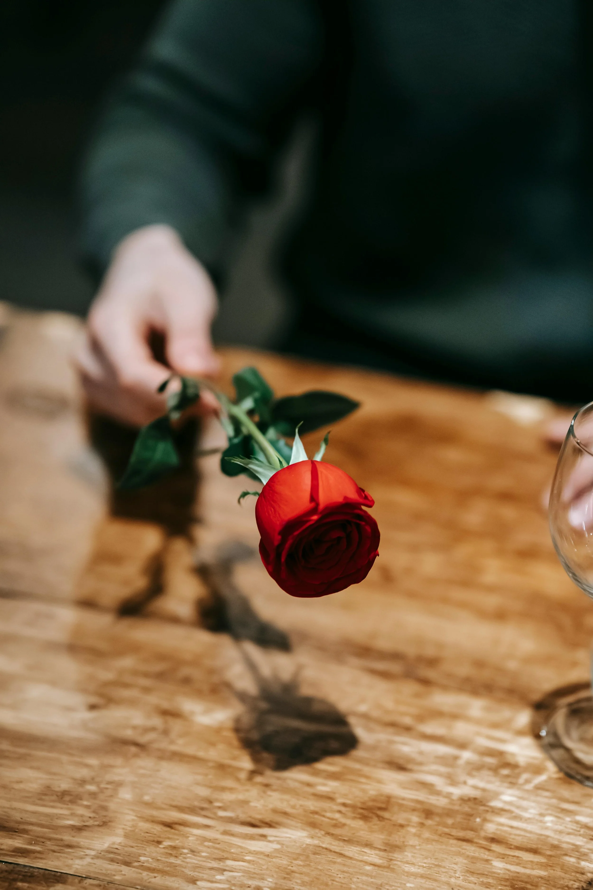 A person's hand holding a single red rose with green leaves over a wooden table, with a glass partially visible on the right. Maria Mountain - Intimacy and Connection Coach. Couples Intimacy Coaching