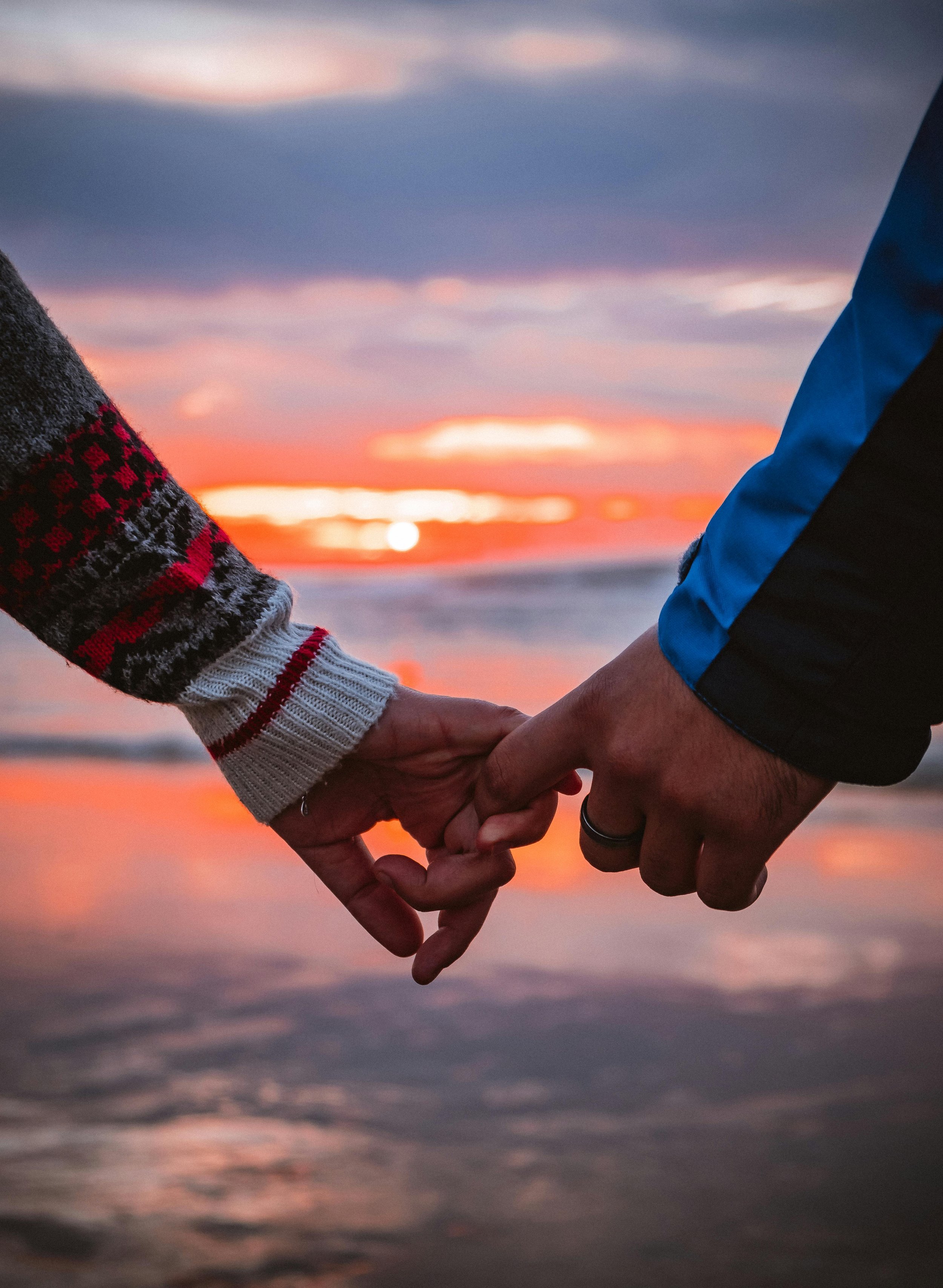 A close-up of two people holding hands on the beach during sunset, with the ocean and colorful sky in the background. Maria Mountain - Intimacy and Connection Coach.